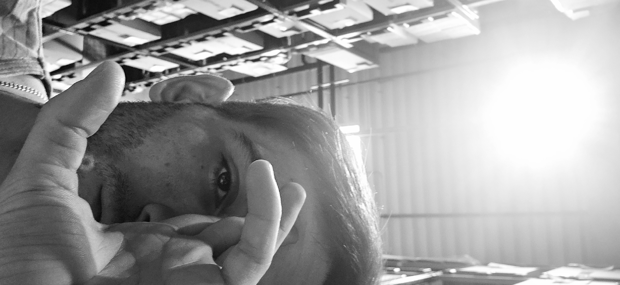 A monochrome photograph shows a person from a low angle, partially obscured by their hand. The person's head is tilted downward, and only one eye is clearly visible. Their hair is swept back. The hand, in the foreground, is positioned near the face. Overhead, a network of suspended rectangular light fixtures or panels is visible, suggesting an indoor environment. In the background, there's a metal wall with vertical lines and a bright light source, which appears to be a window. Due to the lack of specificity in the image, it's not possible to pinpoint an exact location within Plovdiv, Bulgaria. The setting seems to be inside an industrial or commercial building.