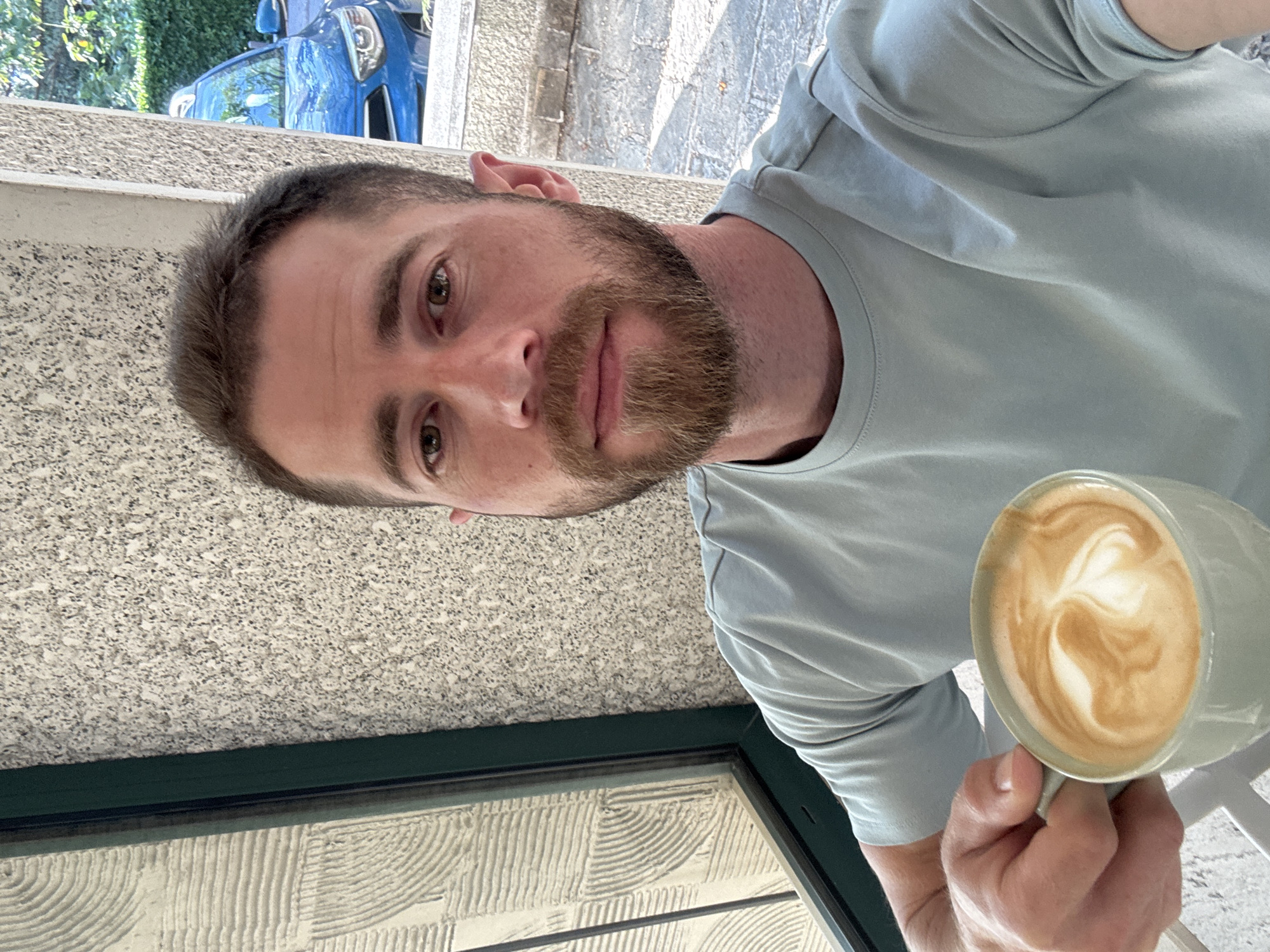 A man with a beard is holding a mug of coffee. The man is wearing a light blue shirt. The photo was taken in Budva, Montenegro, near a window. A car is parked in the background.