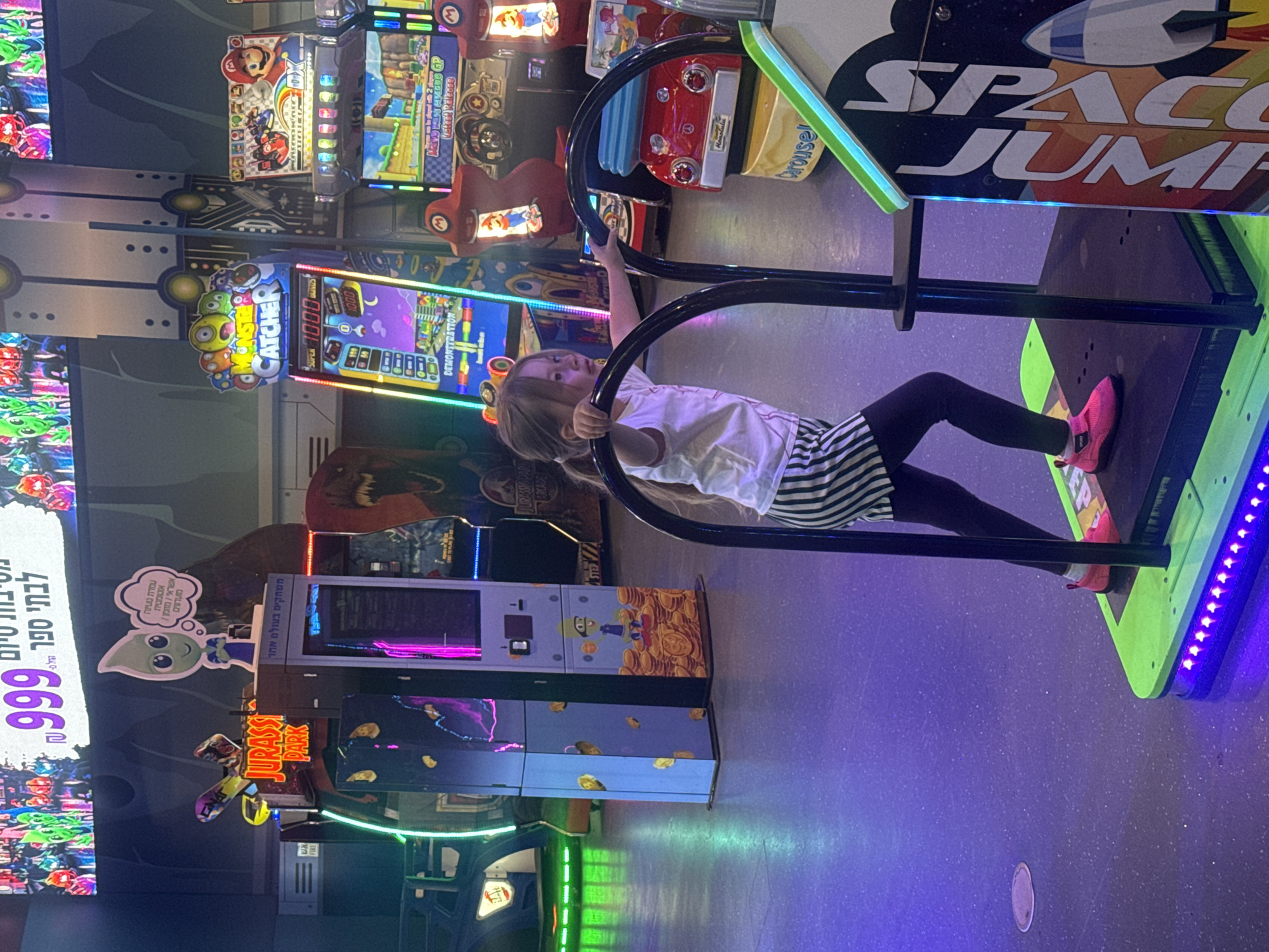 The image captures a young girl playing "Space Jump" at an indoor arcade in Tel-Aviv, Israel. The arcade features various games including "Jurassic Park," "Monster Catcher," and a Mario Kart game. The girl is seen holding onto the safety bar of the "Space Jump" machine while standing on the platform. The arcade has a purple-toned ambiance created by the lighting, and it appears to be located in a shopping center or entertainment complex, identified by the automatic recharge machine.