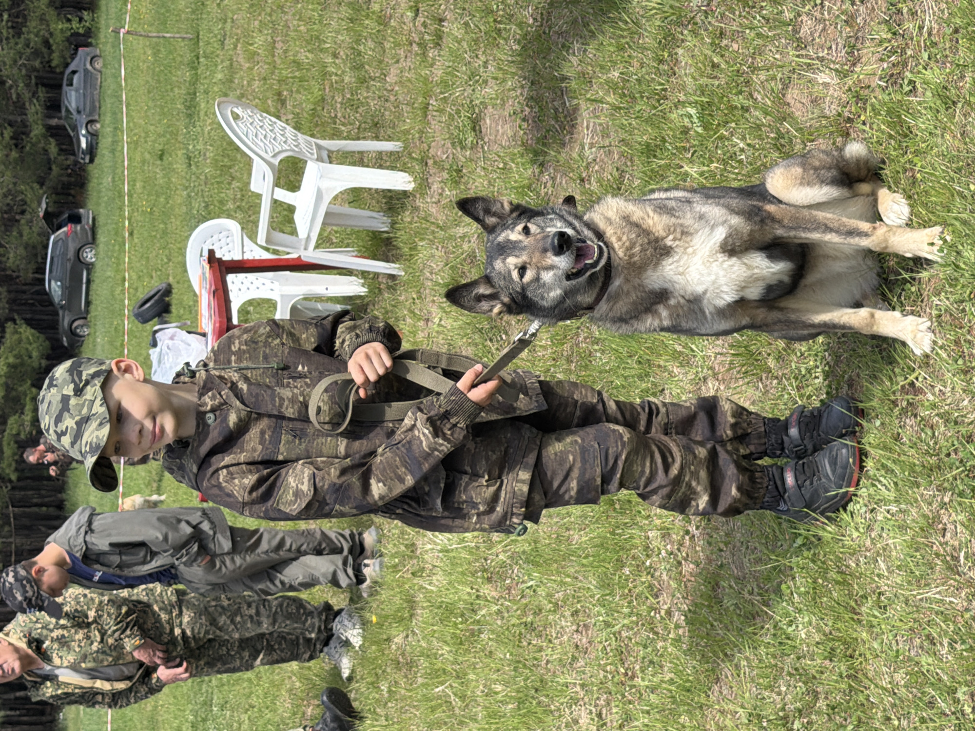 A boy wearing camouflage clothing and a camouflage cap is holding a dog leash. The dog, a medium-sized breed with gray and tan fur, is lying on its back in the grass, appearing relaxed. The boy is standing on a grassy area. Several other people, some also in camouflage, are visible in the background. White plastic chairs and cars are also visible in the background. The location is a grassy field, and the city is Chaikovsky, Russia.
