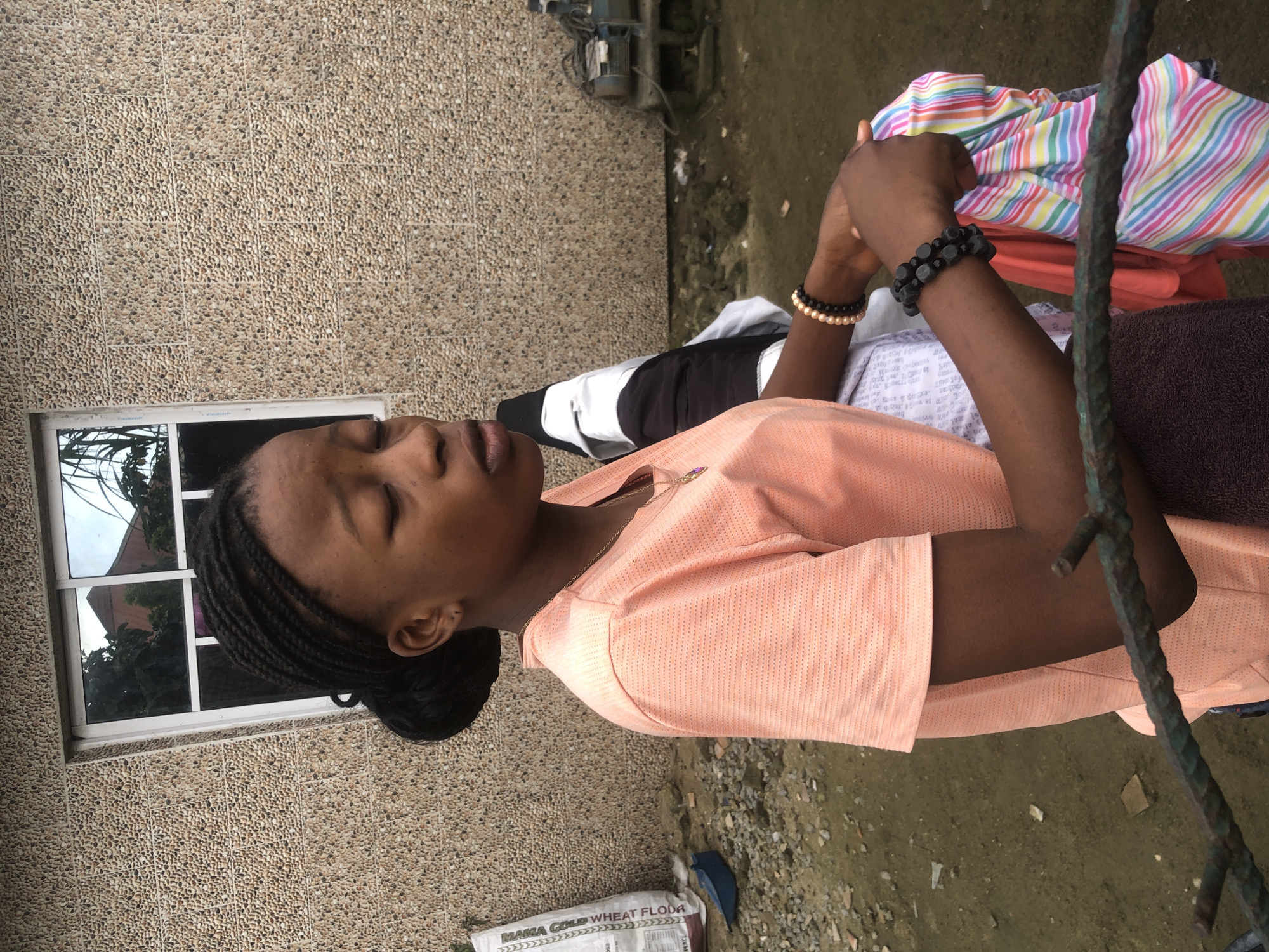 A young woman with dark skin and braided hair is pictured outdoors, facing to the right, with her eyes closed. She is wearing a peach-colored, short-sleeved shirt and a necklace with a pendant. She is holding several folded pieces of colorful, striped fabric in her hands, and has two bracelets on her left wrist. Behind her, a building with a textured, pebble-like facade and a window is visible. To the left of the woman, a white bag labeled "MANGA GOLD WHEAT FLOUR" and a blue object are on the ground, which appears to be dirt and debris. To the right, a piece of machinery, possibly an engine, is situated on a concrete base. The ground is uneven, with patches of mud and scattered rocks. The image appears to have been taken in Ado, Nigeria.