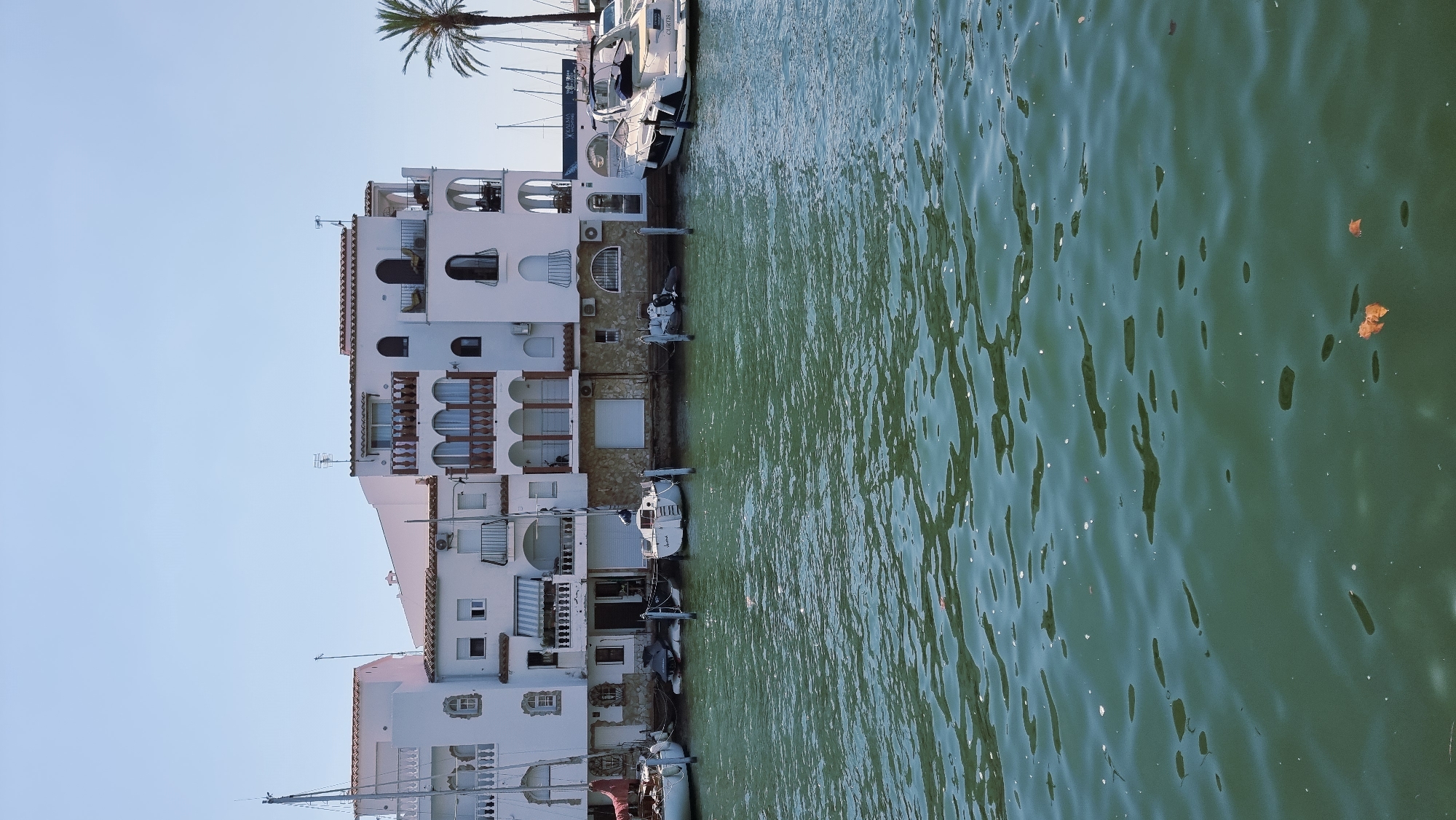 The image shows a waterfront scene in the city of Castelló d'Empúries, Spain. In the foreground, the water of a canal or harbor is a deep green with reflections and some floating debris. Several boats are docked along the quay. To the left, a white sailboat with a red sail is partially visible. Next to it is another white sailboat. In the center, a small white sailboat with the name "June" on its stern is moored. To the right of this, a small dinghy or inflatable boat is tied up. Further to the right, a larger white motor yacht named "Curtis" is docked.
Behind the boats, a row of white buildings lines the waterfront. These buildings have multiple stories, balconies with arches, and red-tiled roofs. Some of the buildings feature arched windows and doorways. There are air conditioning units visible on some of the building facades. A palm tree is visible behind the buildings on the right side of the image. The sky is clear and blue. The overall impression is a sunny day in a picturesque Mediterranean marina.