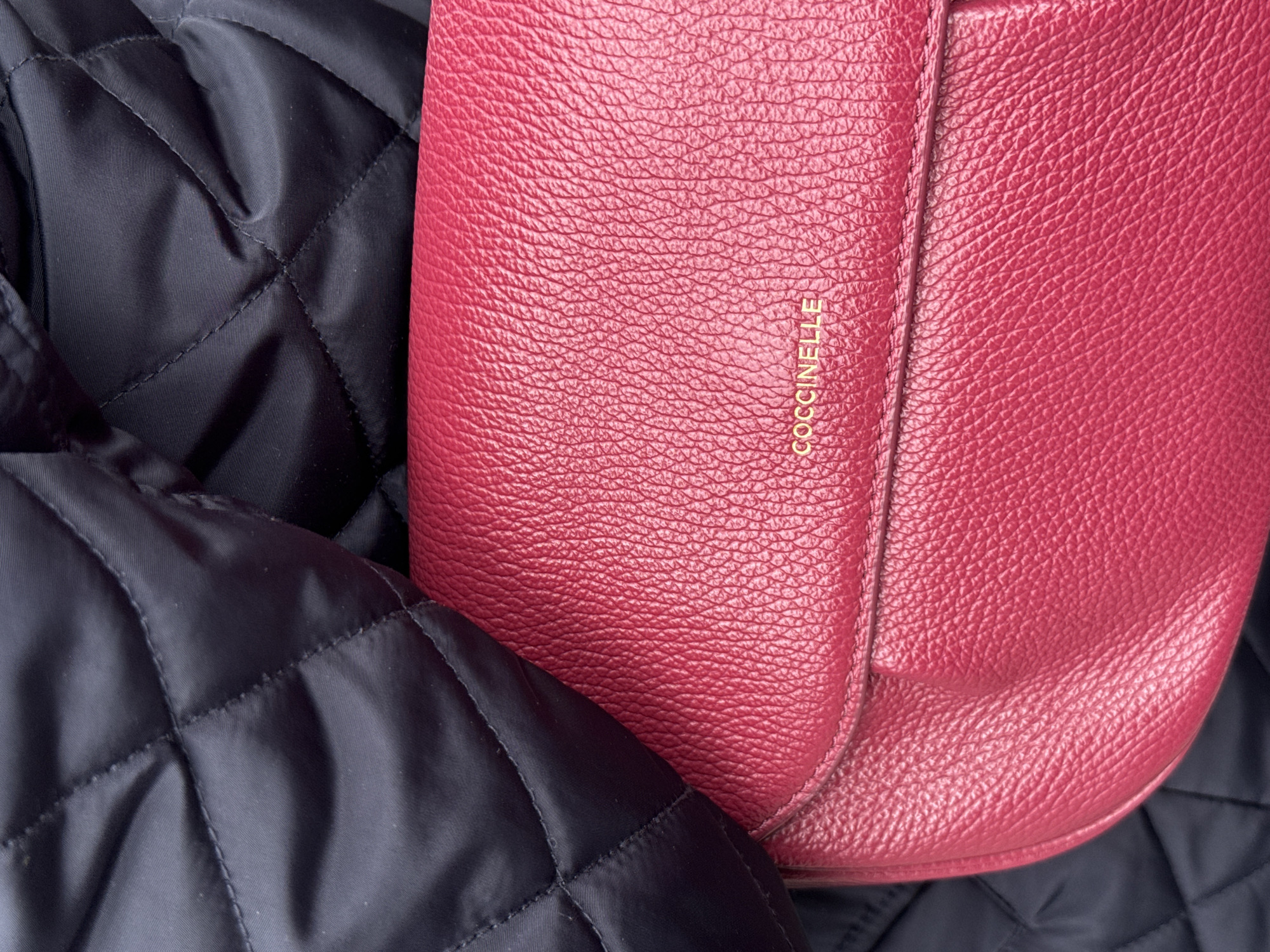 A close-up shot shows a red Coccinelle leather handbag resting against a black, quilted fabric. The handbag features a flap closure and the brand name is embossed in gold lettering. The quilted fabric has a diamond pattern and appears to be part of a jacket. The scene is indoors with controlled lighting. There is no evidence to suggest that it is in Paris, France.