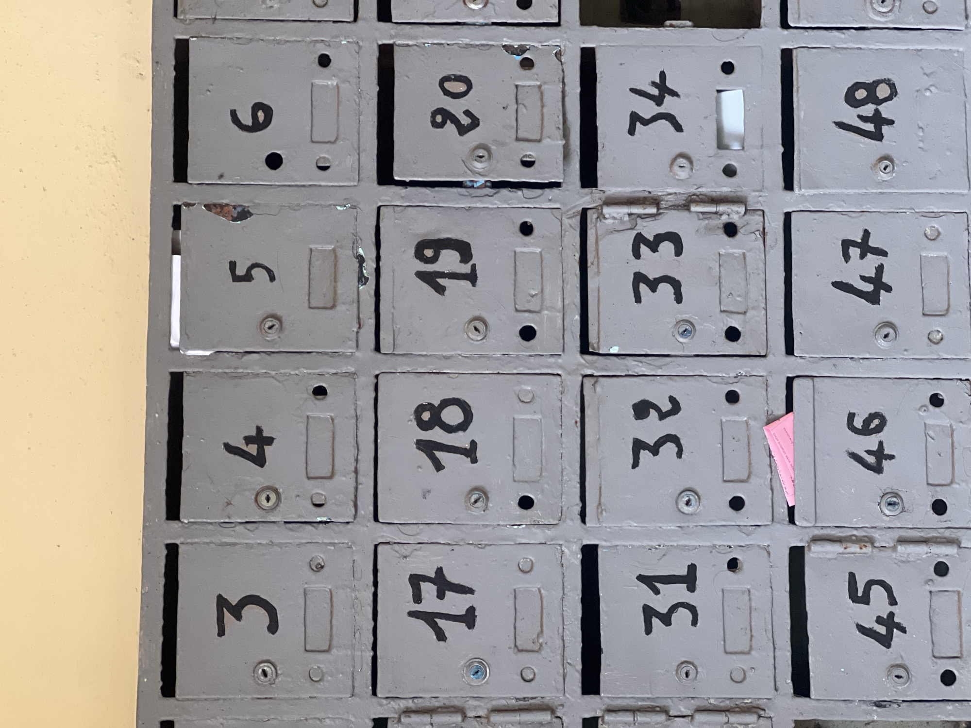 The image displays a close-up, slightly overhead view of an old, wall-mounted bank of grey metal mailboxes. The mailboxes are arranged in a grid, showing four columns and four visible rows, set against a plain, light yellow wall to the left. Each mailbox door features a hand-painted black number, a keyhole or lock mechanism, and a small rectangular slot.
The numbers on the mailboxes descend numerically within each column. From left to right, the visible numbering pattern is:
* Column 1: 6, 5, 4, 3
* Column 2: 20, 19, 18, 17
* Column 3: 34, 33, 32, 31
* Column 4: 48, 47, 46, 45
The mailboxes appear well-worn, with chipped paint, scuffs, and some visible rust, indicating age and frequent use. Some of the mailbox doors are slightly ajar, and a small piece of white paper is visible inside mailbox 34. A distinct pink piece of paper is seen sticking out from the opening of mailbox 46, suggesting recent mail delivery. The scene depicts a functional, communal mailbox system, typical of apartment buildings or older residential structures. No people are present, and the overall impression is that of an aged but active mail collection point in a location such as Casablanca, Morocco.