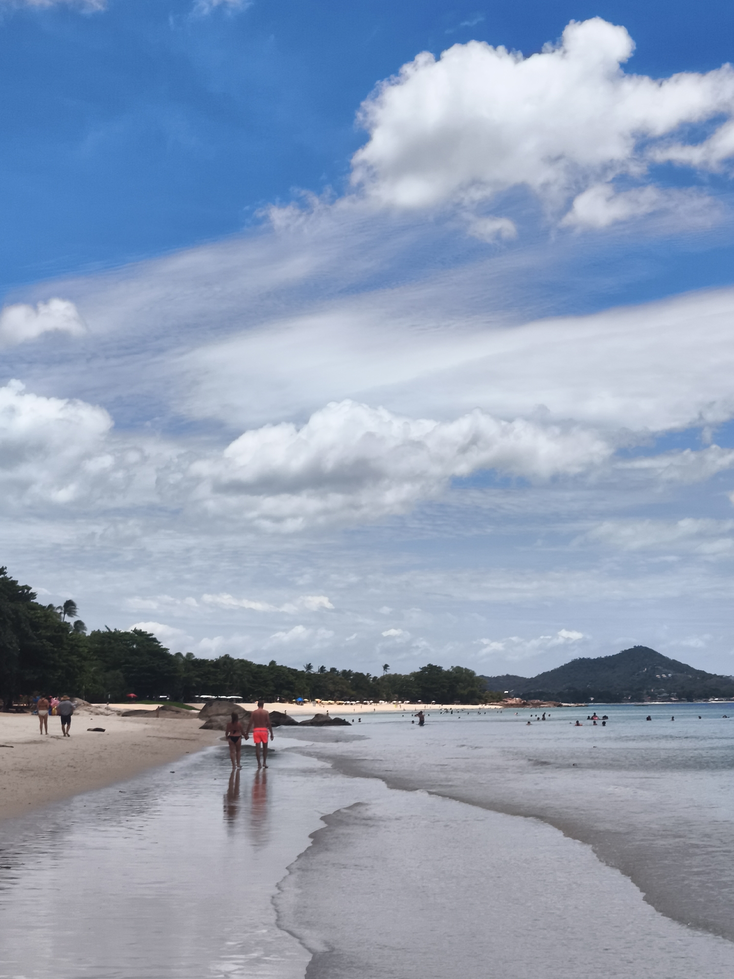 The image depicts a tropical beach scene in the Ko Samui District of Thailand. The foreground features the wet sand of the shoreline, with gentle waves lapping at the edge, creating reflections of the sky and people. In the mid-ground, a group of people are walking along the beach and in the shallow water. Closer to the viewer, a couple in swimsuits walks hand-in-hand, their reflections visible in the wet sand. Further down the beach, several other individuals and groups are visible, some wading in the water, others walking on the sand, and some gathered under umbrellas. The background shows a line of lush green trees and vegetation, followed by a hilly, mountainous landscape under a vast, partly cloudy sky. The overall impression is a lively, sunny day at a popular beach resort.