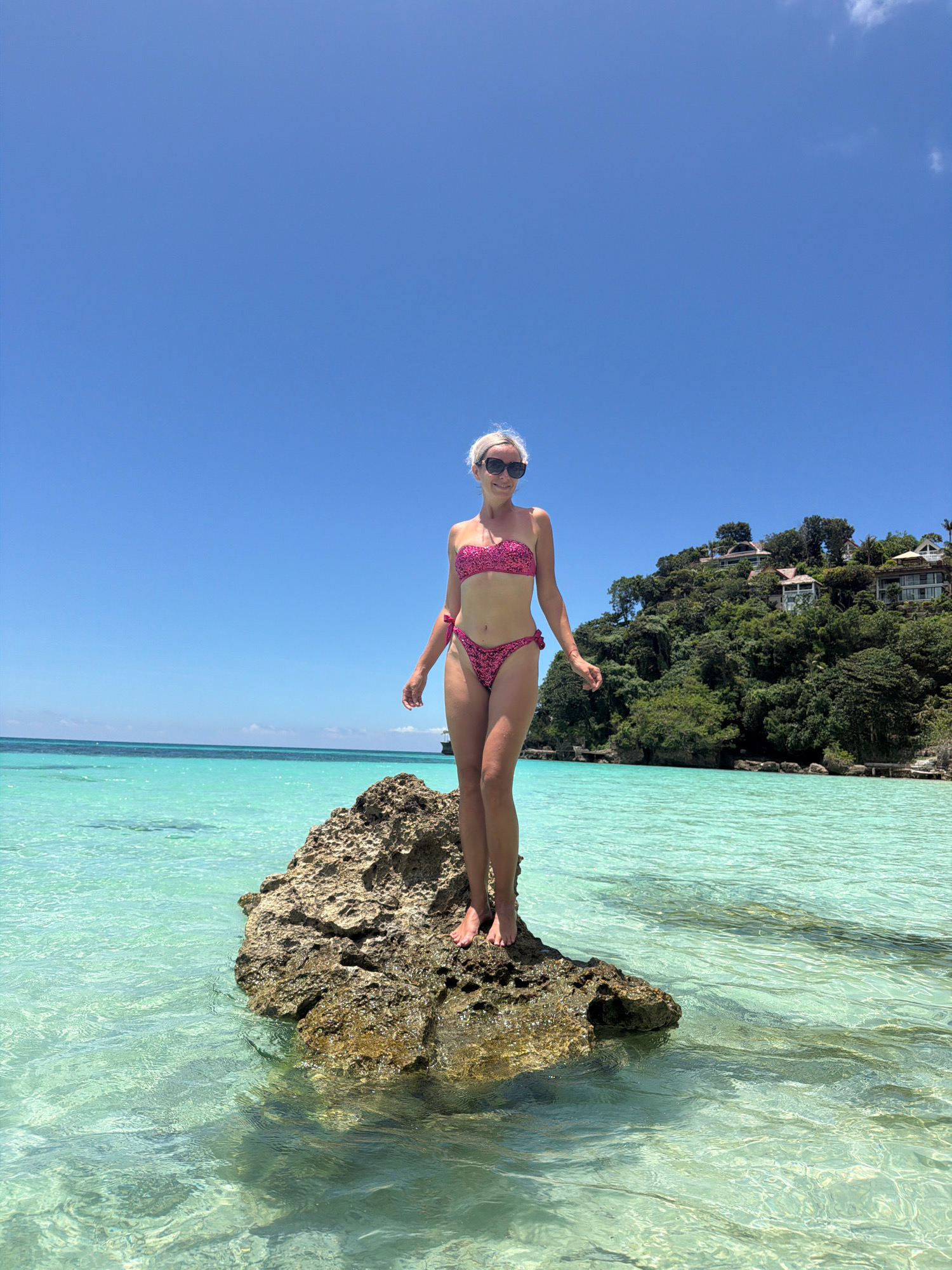 A woman stands on a rock in the shallow, clear turquoise water of a tropical beach. She is wearing a pink bikini with a black leopard print and sunglasses. Behind her, a lush green hillside is dotted with white houses. The sky is a bright, cloudless blue. This scene appears to be at Station 1, Boracay Island, Malay, Philippines.