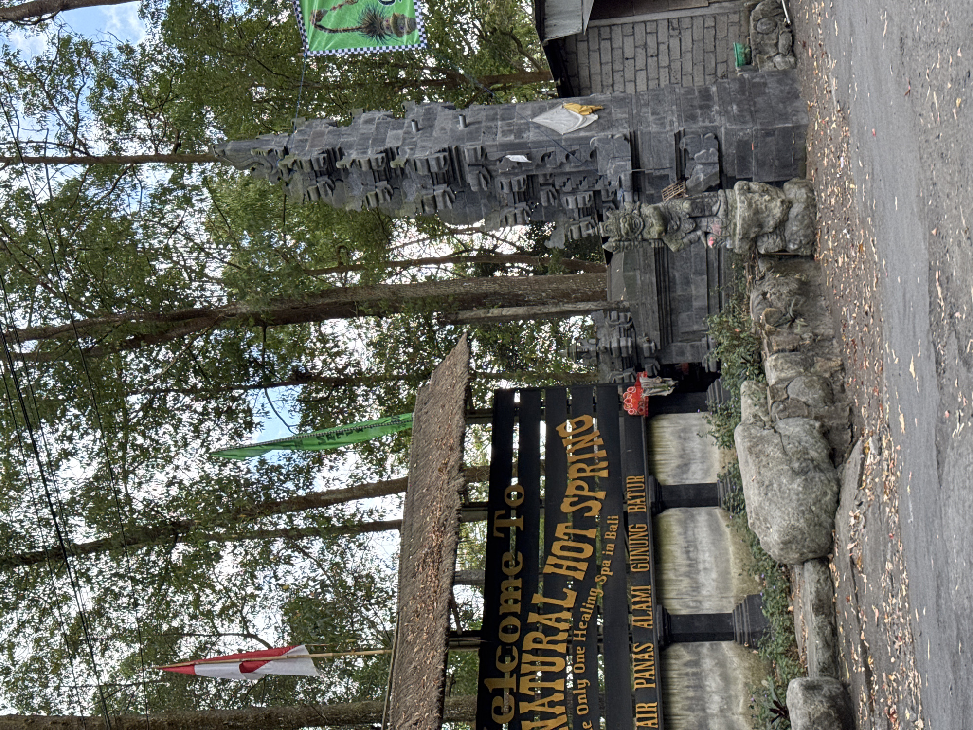 The image shows the entrance to a natural hot spring in Kintamani, Indonesia, likely Batur Natural Hot Spring. A large, dark sign with yellow lettering reads "Welcome To NATURAL HOT SPRING" and below it, "The Only One Healing Spa in Bali" and "AIR PANAS ALAMI GUNUNG BATUR" (Natural Hot Spring Mount Batur). The sign is framed by a thatched roof structure. To the right of the sign is a traditional Balinese stone gate, intricately carved and weathered. A small statue of a deity or guardian figure stands at the base of the gate. Indonesian flags, red and white, are visible on poles near the sign, and a green banner hangs from the gate structure. The area is surrounded by tall trees with lush green foliage. The ground is paved with asphalt and littered with fallen leaves. The entrance appears to be a natural, rustic setting.