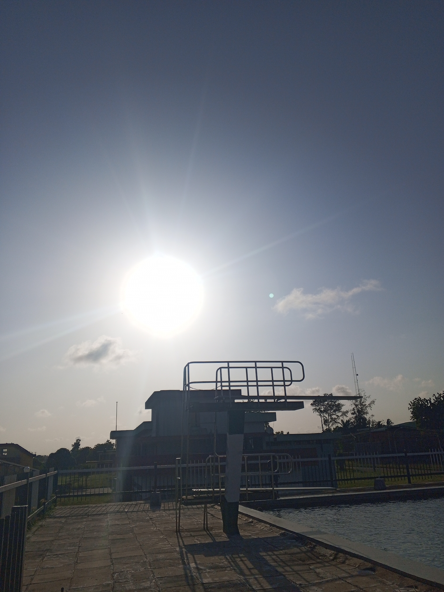 The image depicts an outdoor swimming pool in Tawpo, Nigeria, under a bright, high sun. A diving board is prominent in the foreground, positioned on a tiled surface adjacent to the pool. A low metal fence surrounds the pool area, separating it from a grassy area and some trees. In the background, there is a multi-story building. The sky is clear with a few scattered clouds. No people are visible.