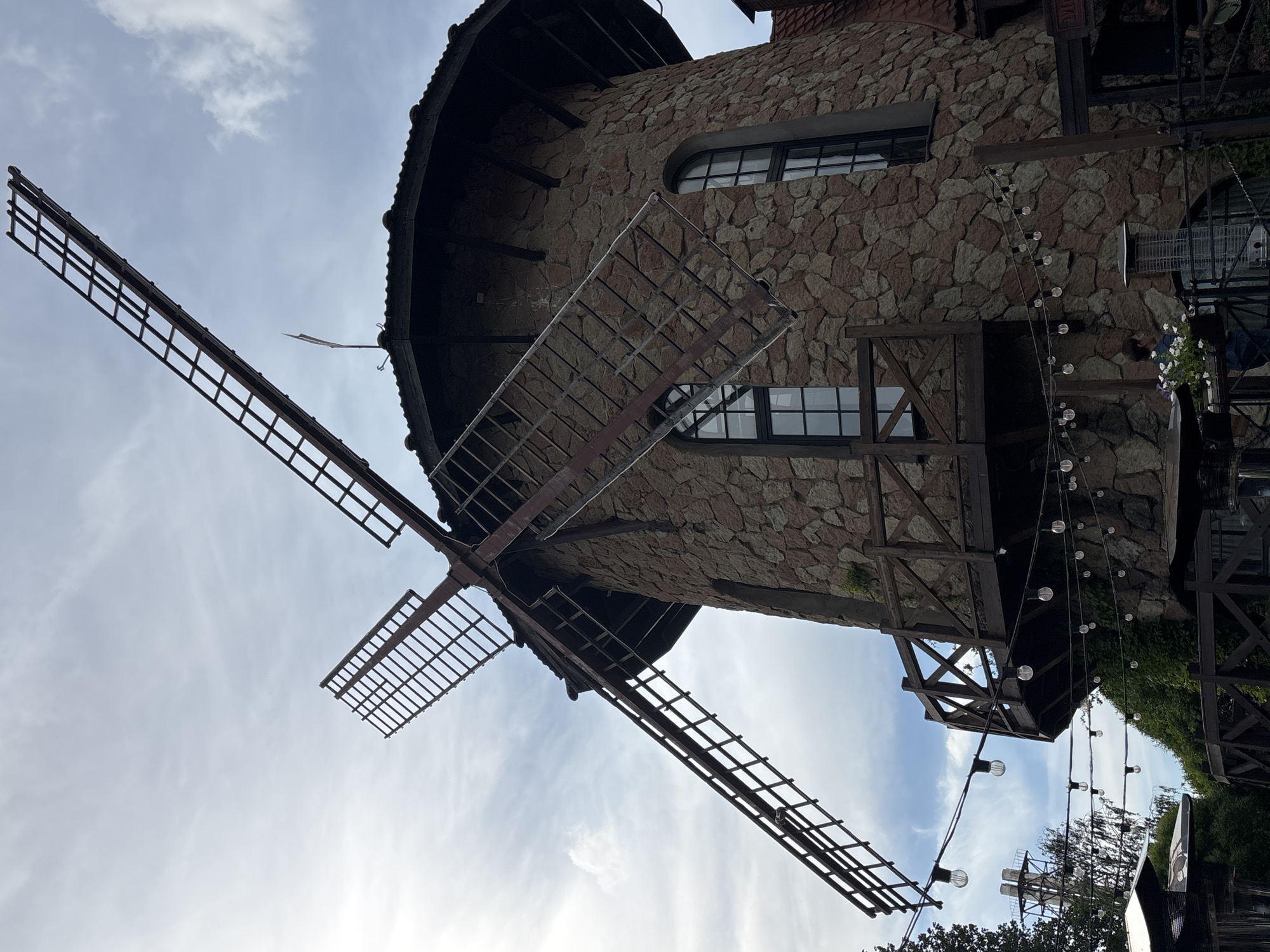 An architectural shot captures a stone-built structure resembling a windmill, likely located in Saint Petersburg, Russia. The building's cylindrical body is constructed from varied, earthy-toned stone blocks. Protruding from the top section are four dark, latticed windmill blades, two of which are prominently visible, extending outwards and downwards against the sky. The building features dark-framed windows, including one distinct arched window and another rectangular one, both divided into multiple panes.
A rustic wooden balcony or decorative structure is attached to the stone facade on the right side. Below this, a string of round, decorative lights hangs vertically, suggesting an outdoor patio or dining area. Lush green foliage, possibly ivy or a climbing plant, adorns parts of the building's right side, adding to its picturesque appeal. The scene unfolds under a bright, partly cloudy sky, indicating daytime with a soft, diffused light, possibly during the morning or late afternoon. No people are visible, and there is no discernible text on the building or surrounding elements. The structure appears to be a unique commercial establishment, perhaps a restaurant or cafe, designed with a traditional European windmill aesthetic.
