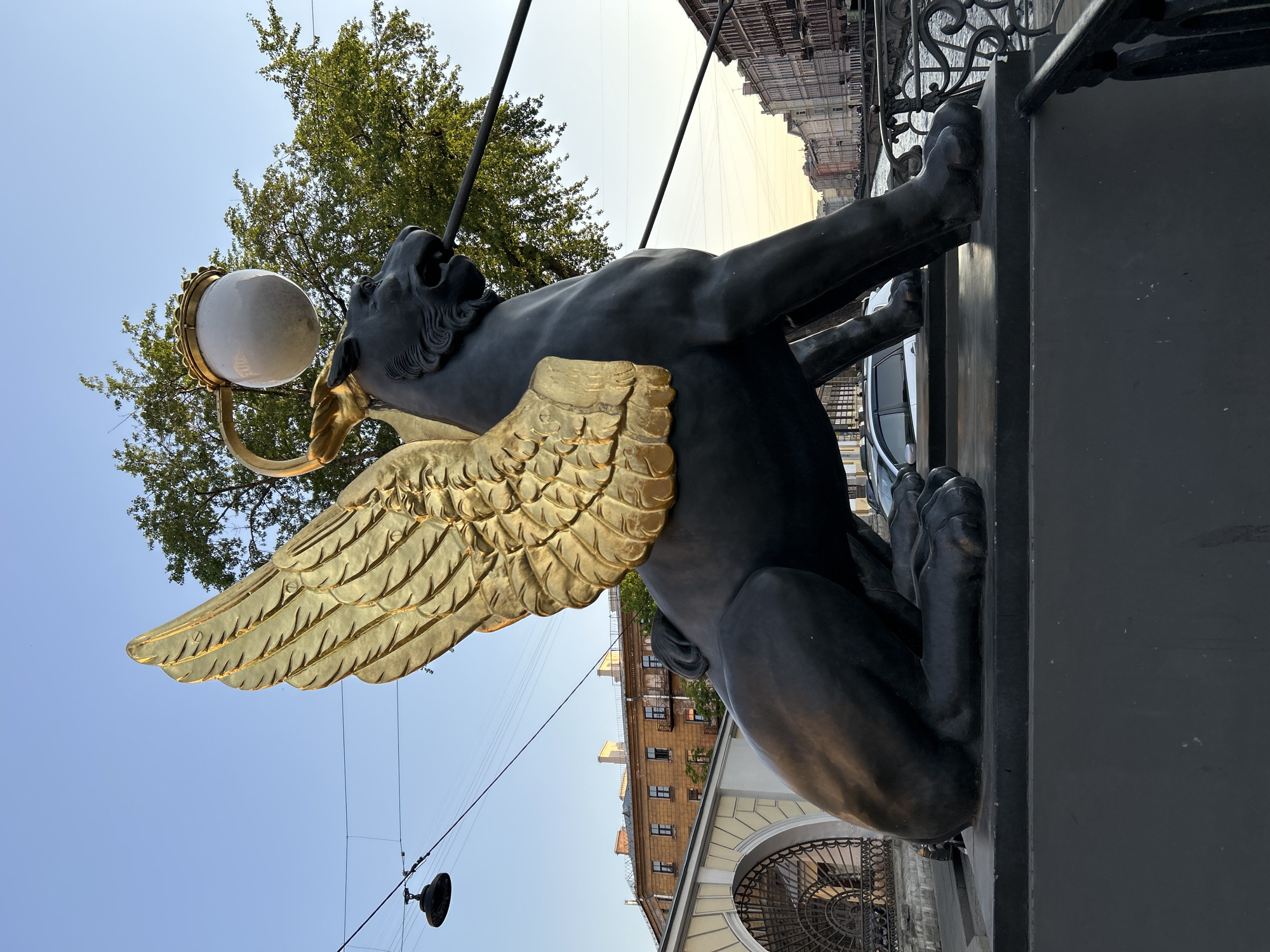 The image shows a winged griffin statue from the Bank Bridge in Saint Petersburg, Russia. The statue is made of dark metal with gilded wings. It holds a white spherical lamp in its mouth. In the background, there are buildings with windows and architectural details, as well as a tree with green leaves. The sky is light blue. Power lines with a round streetlight are visible in the lower left corner. The statue is mounted on a dark-colored pedestal or railing. Part of a vehicle is visible in the background near the upper right. An arched gate with decorative ironwork is also partially visible.