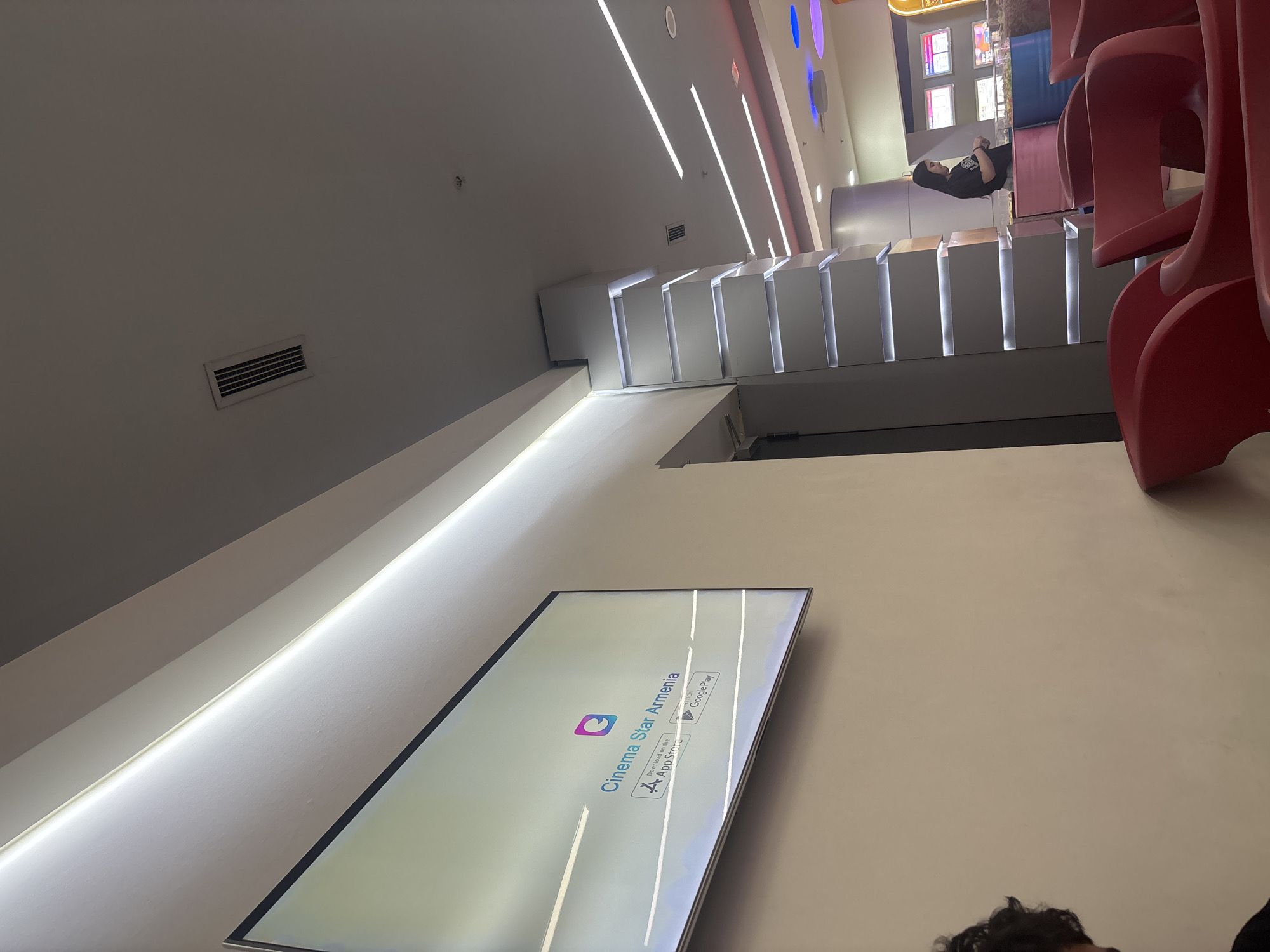 This image captures a modern indoor waiting area, likely within a cinema lobby in Yerevan, Armenia. The scene is brightly illuminated by linear ceiling lights and integrated lighting within architectural features.
In the midground, a prominent flat-screen display is mounted on a light-colored wall, showing the text "Cinema Star Armenia" with icons for "Download on the App Store" and "GET IT ON Google Play," indicating a mobile application. To the right of the screen, a contemporary architectural feature consists of several ascending white steps or platforms, each with internal illumination. Further to the right, a stack of several distinctive red, high-backed molded chairs is partially visible, suggesting a seating area.
In the upper right background, a person with dark hair, wearing a dark top, is seated and appears to be engaged with a mobile phone. The top of another person's head, featuring dark, curly hair, is visible in the lower right foreground. The background also reveals a service counter or concession area with display screens and blue and purple accent lighting. A ventilation grille is visible on the ceiling. The overall setting is clean, modern, and suggests a public entertainment venue.