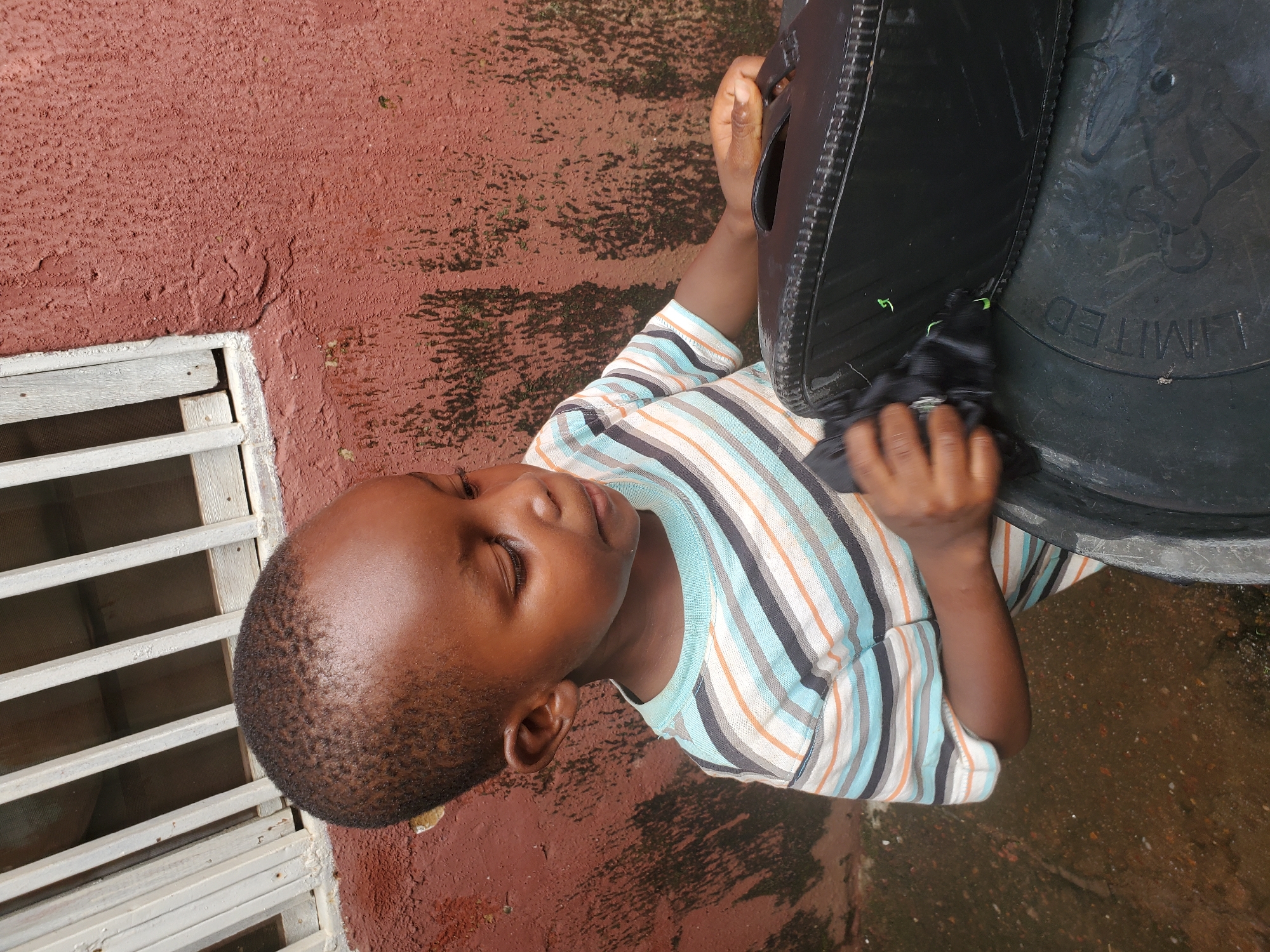 A young Black boy, likely under the age of six, is focused on cleaning a black object with a dark cloth. He is wearing a horizontally striped t-shirt with bands of white, blue, black, and orange. The boy is positioned in front of a reddish-brown textured wall, which appears weathered and has some green moss or algae growing on it. To the left of the wall, there's a white barred window frame. The ground beneath the boy is wet and appears to be composed of dirt and some debris. In the foreground, the boy is touching a black, rectangular object, possibly a bag or case, which is resting on a larger black cylindrical container. The setting suggests an outdoor, possibly a domestic, environment in Jos, Nigeria.