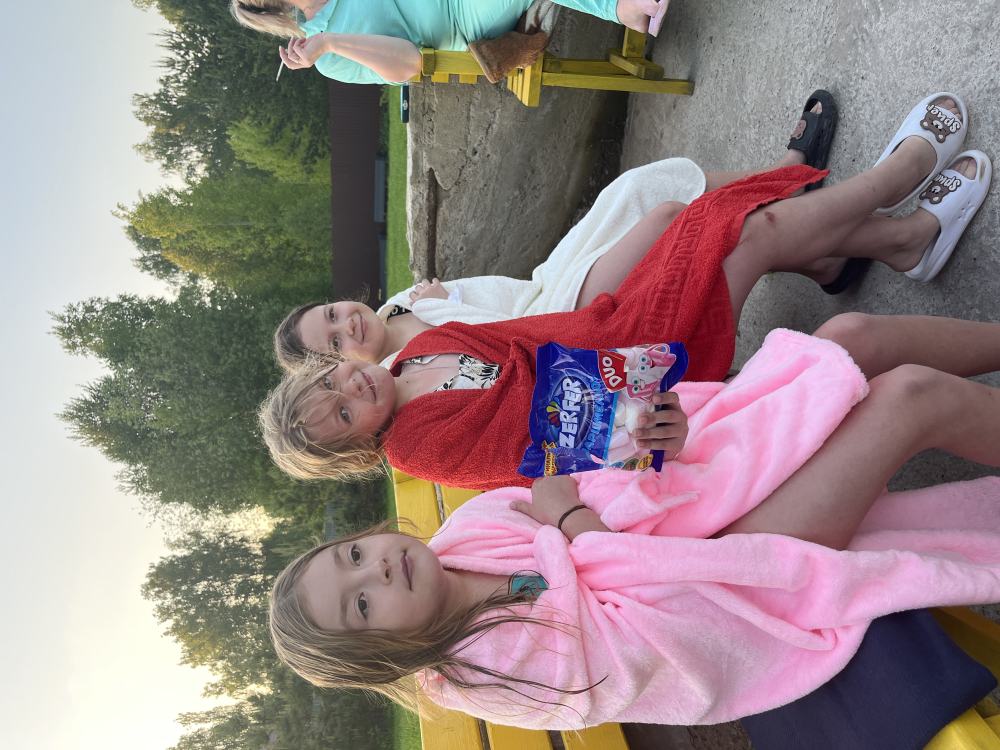 Three children are sitting on a yellow bench, wrapped in bathrobes: a girl in pink in the foreground, a girl in red with a package of Zerfer Duo marshmallows in the middle, and a girl in white in the background. An adult is visible on another bench in the upper left of the frame. The location is outdoor with trees and building visible in the background, Новая Чернушка, Russia.