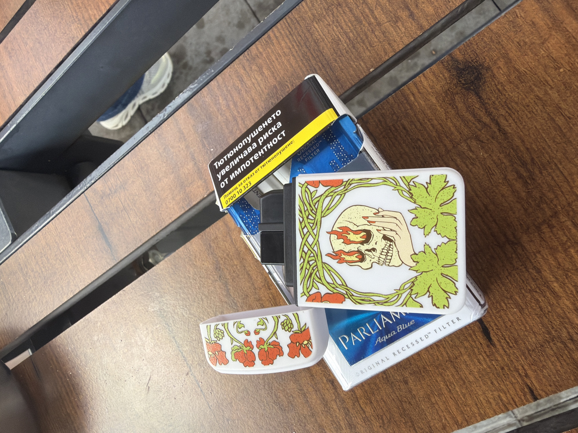 The image presents an overhead view of a pack of cigarettes and an electronic cigarette device situated on a dark brown wooden slatted surface in Osikovitsa, Bulgaria. The cigarette pack, visible in blue and white, is identified as "Parliament Aqua Blue" with an "ORIGINAL RECESSED™ FILTER". A yellow and black health warning label in Bulgarian reads "Тютюнопушенето увеличава риска от импотентност" (Smoking increases the risk of impotence), accompanied by a helpline number "0700 10 323". Resting on the cigarette pack is an open white electronic cigarette or vape device. Its hinged lid displays a repeating pattern of red flowers and green foliage. The main body of the device features a central graphic of a skull with prominent flaming eye sockets, partially held by a hand with red fingernails, encircled by green leafy vines. In the background, a dark metal structure divides the wooden surface. Through an opening in this structure, a person's foot wearing a white and blue athletic shoe is visible on a grey ground surface.