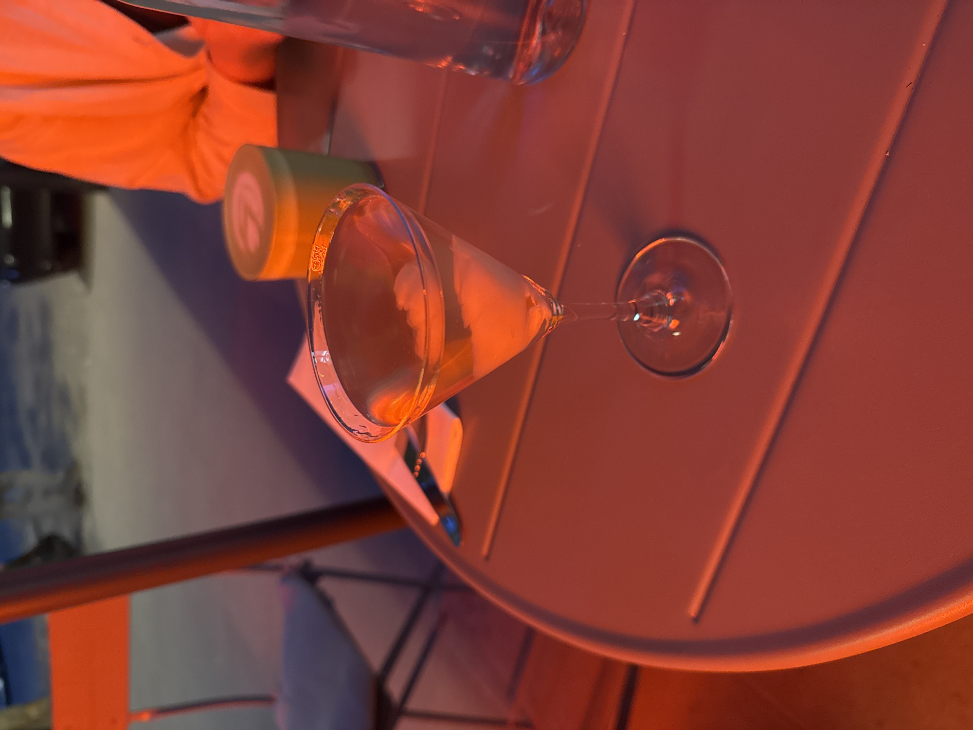 The image shows a cocktail on a red-orange table, likely outdoors, with a green container nearby. A person's arm in an orange jacket is visible to the side. Another beverage in a clear glass is present. The cocktail appears to be a martini or similar drink in a stemmed glass. The environment is illuminated with warm-toned lighting. There is an indiscernible background visible in the lower left, suggesting an urban setting. However, there is not enough information to definitively confirm the location as Warsaw, Poland, or to provide precise details about actions, interactions, and other people in the scene.