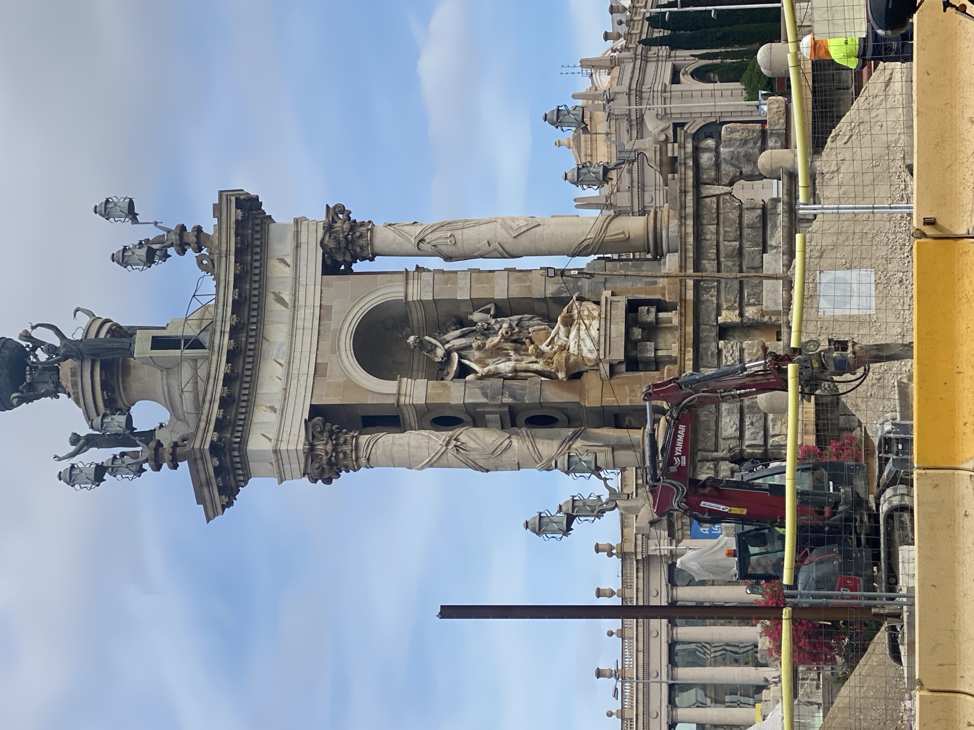 The image depicts a construction site in front of a grand monument in Barcelona, Spain, specifically the Venetian Towers, located at the entrance to the Montjuïc exhibition site. In the foreground, a red and black Yanmar excavator is positioned behind a yellow construction barrier. Construction materials, including dirt and sand, are visible. A construction worker wearing a yellow safety vest and an orange hard hat is standing near the barrier, facing away from the camera.
The central focus of the image is the monument, characterized by its intricate classical architecture. It features ornate columns, statues, and multiple lamp posts with decorative lanterns. The monument appears to be undergoing some form of renovation or construction, as suggested by the presence of the machinery and barriers. The sky above is partly cloudy, indicating daylight. Behind the monument, another large, classical building with arched windows and colonnades is partially visible, suggesting a larger architectural complex. Lush green trees are seen along the right side of the background.