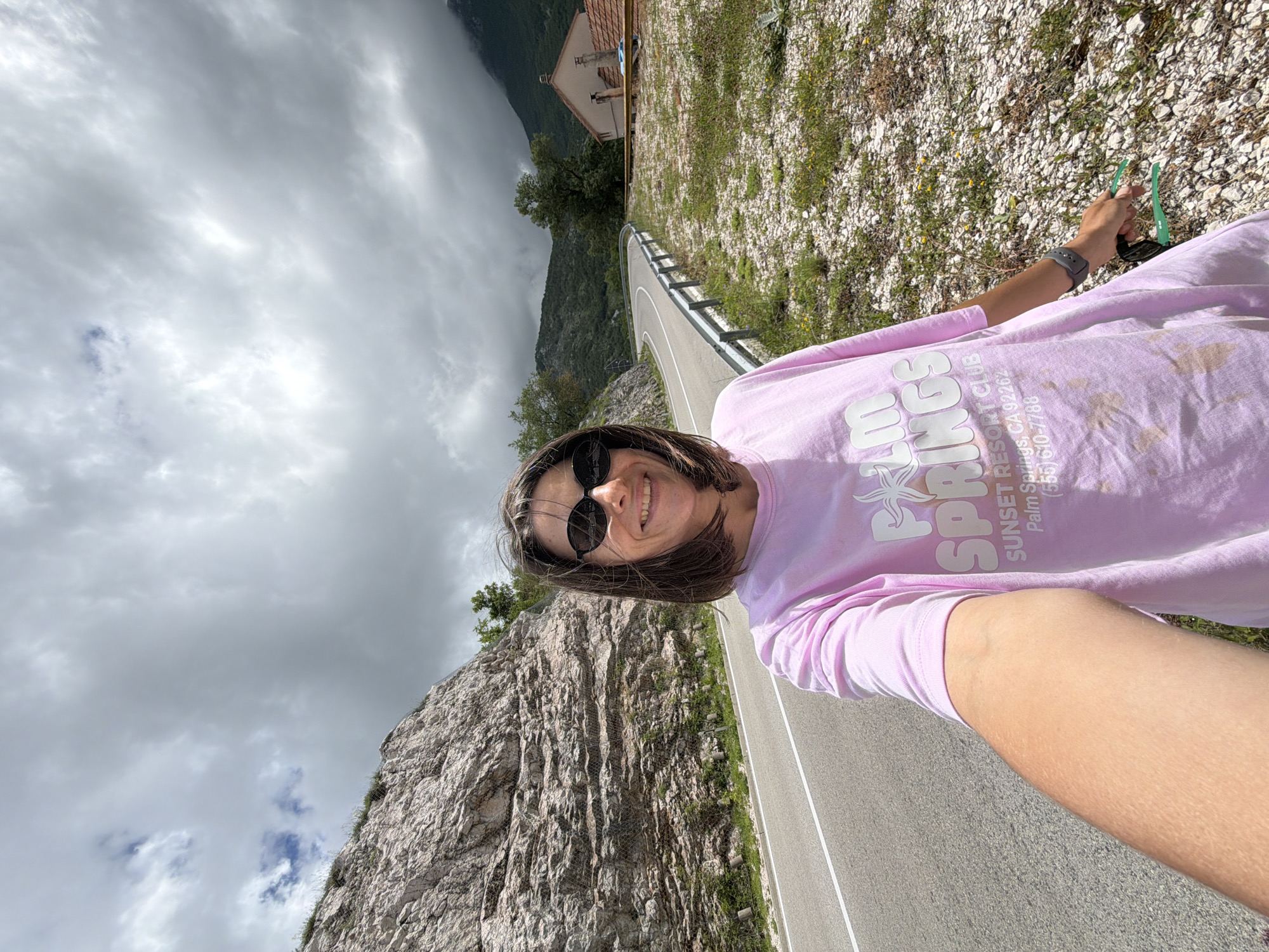The image displays a selfie taken by an individual, likely female, standing on the side of a winding mountain road. The person is smiling and wearing dark sunglasses, dressed in a light pink long-sleeved t-shirt. A watch is visible on her left wrist, and she holds a pair of green-framed sunglasses in her right hand.
The background features a dramatic mountainous landscape. To the left, a large, rugged rocky mountainside rises steeply, exhibiting visible rock layers and sparse vegetation, with some areas showing what appears to be rockfall netting. The asphalt road curves sharply, marked with a white line and bordered by a metal guardrail on the right. Beyond the guardrail, the terrain slopes down, covered in light-colored rocks, grass, and small plants. In the upper background, amidst darker, tree-covered peaks, a small building with a reddish-brown tiled roof is visible. The sky is predominantly overcast with gray clouds, though patches of blue are discernible.
The text on the pink t-shirt clearly reads "PALM SPRINGS SUNSET RESORT CLUB CA 92262 (555) 610-7788". The scene depicts a daytime moment in a scenic, mountainous area, consistent with Žanjev Do, Montenegro. The individual's smile suggests a pleasant experience.