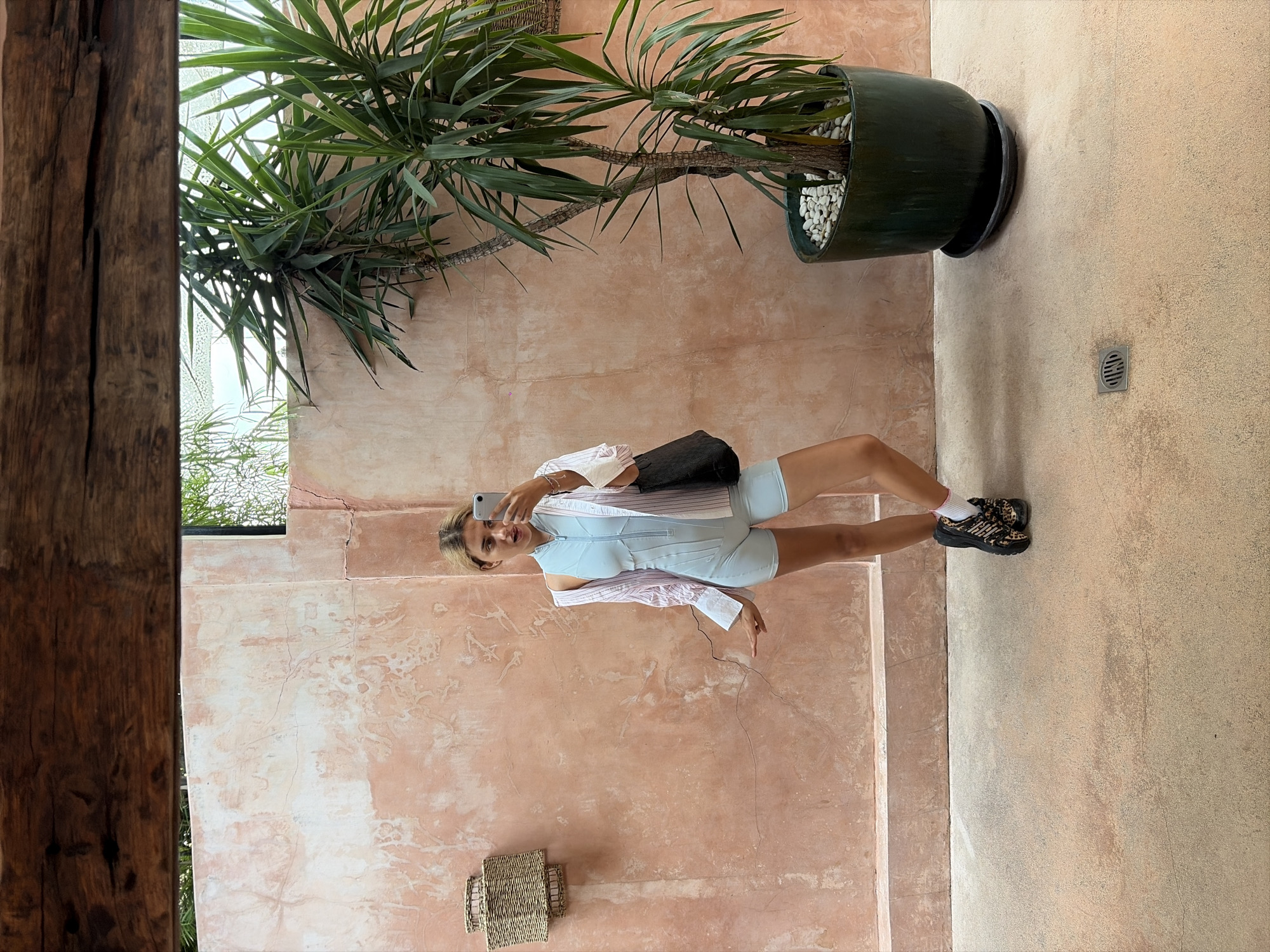 A young woman stands facing a mirror in what appears to be an outdoor or semi-outdoor setting. She is taking a selfie with her phone. She is wearing a light blue one-piece bodysuit with a zipper down the front, a striped pink and white open shirt over it, white socks, and black and leopard print platform shoes. She has a dark tote bag slung over her right shoulder.
Behind her, a salmon-pink textured wall is visible, with a woven light fixture mounted on it to the left. To the right of the woman, a large potted yucca plant stands. The ground is a light concrete surface with some visible cracks. The overall environment suggests a warm climate, possibly a resort or stylish outdoor area in North Kuta, Indonesia, given the vegetation and architectural style. The lighting indicates it is daytime.