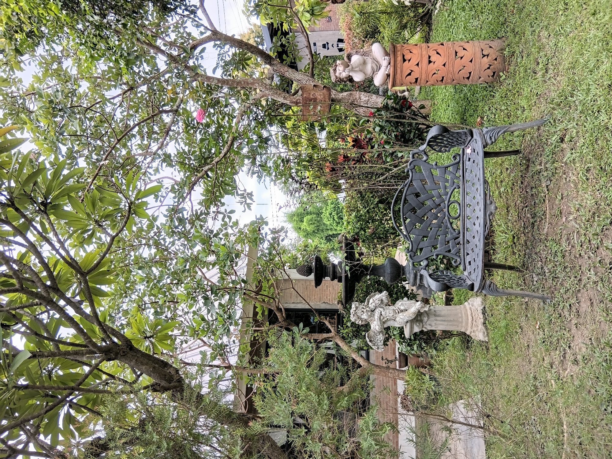 In the city of Ban Tong, Thailand, a tranquil garden scene unfolds. A black wrought-iron bench sits centrally on a patch of sparse grass. To the left, a white stone statue of an angel with outstretched wings stands on a pedestal. Behind the bench, a dark, tiered fountain is visible. To the right of the bench, another white stone statue depicts a cherubic figure, seated and holding a small bird, placed atop a decorative terracotta pillar. Lush green foliage and trees surround the area, with a building constructed of brick visible in the background. The sky above is partially obscured by the branches of a large tree.