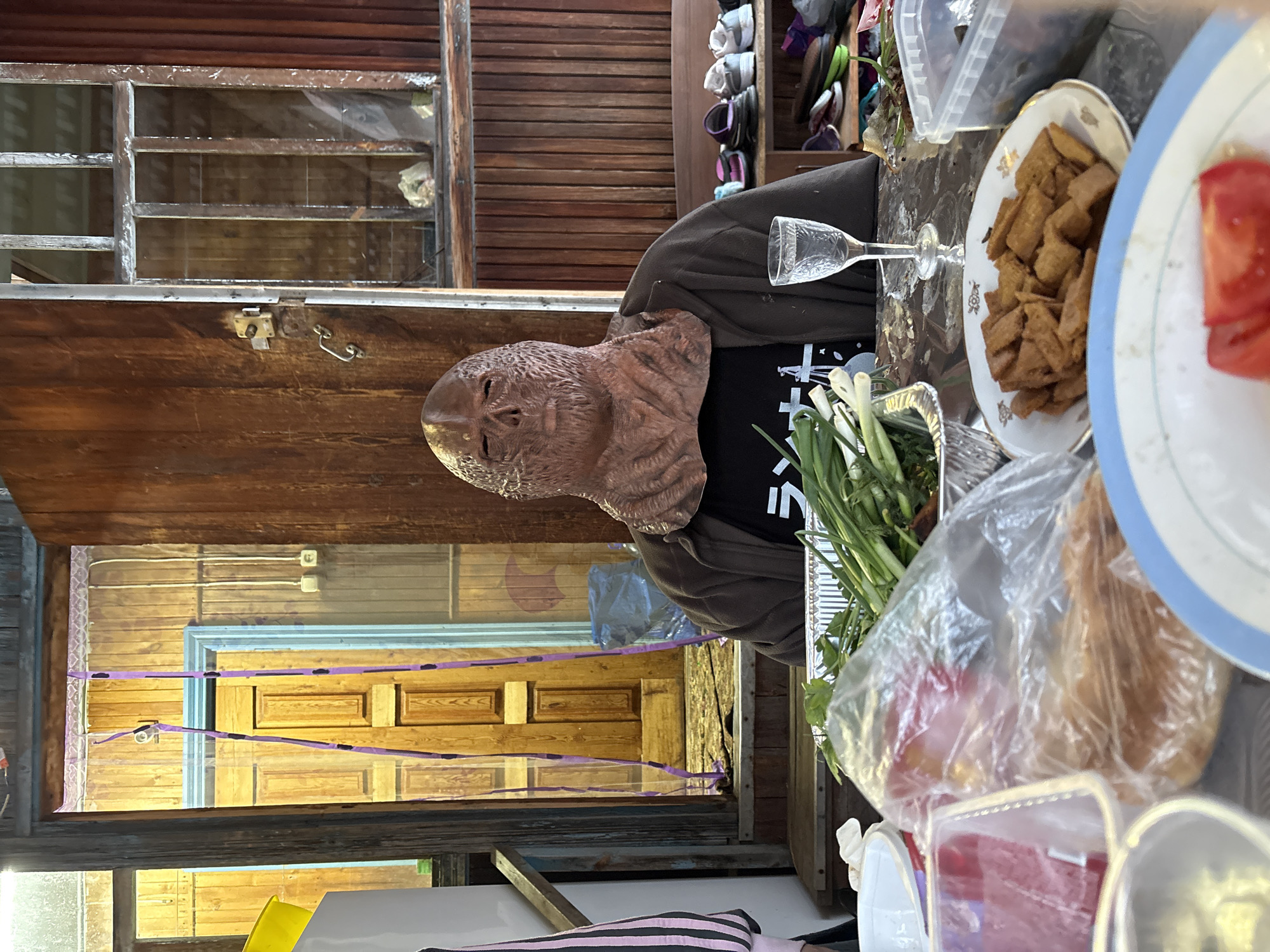 A person wearing a monster mask is seated at a table laden with food and drinks, seemingly in an outdoor setting, given the surrounding wooden structures and vegetation. The table includes items such as a glass of what appears to be a clear liquid, plates with fried food and sliced tomatoes, and packages of fresh vegetables. The backdrop consists of a wooden wall with a small window, and a partial view into a room with a wooden door decorated with purple accents. This image was taken in the city of Zheleznodorozhny, Russia.