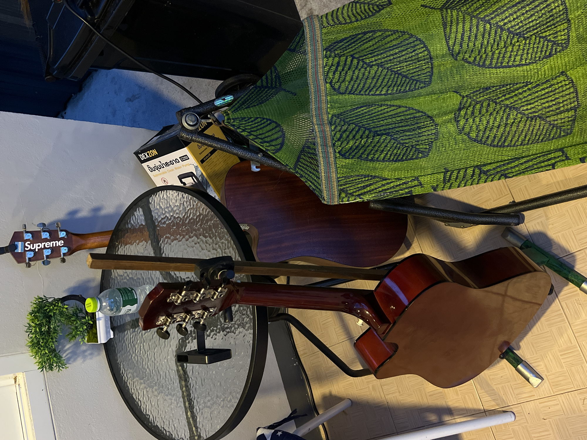 The image depicts a cluttered scene of objects in a state of disarray. Two acoustic guitars are visible: one has a "Supreme" label near the tuning pegs, and the other is dark red-brown. These guitars rest on or near a round glass-topped table. There is a green leafy object in a white holder with a water bottle on the table. A yellow box with Thai writing on it, labeled "DEXZON", is partially visible. A green hammock is partially visible near the top right. The scene takes place on a light-colored tiled surface. A dark object, likely a piece of furniture or equipment, is visible in the upper left corner.