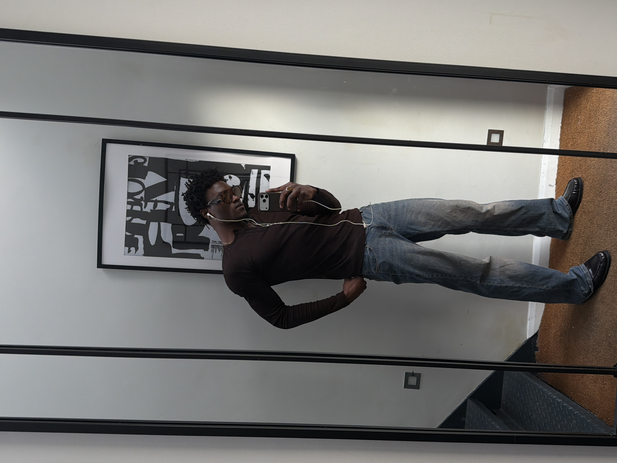 A person is taking a selfie in a mirror. The person is wearing glasses, a brown long-sleeve shirt, blue jeans, and black shoes. They have headphones in. Behind them is a framed black and white abstract print hanging on a white wall. A small square light switch is also visible on the wall. The reflection in the mirror also shows a staircase with metal steps and a brown carpeted floor. The location is London, United Kingdom.