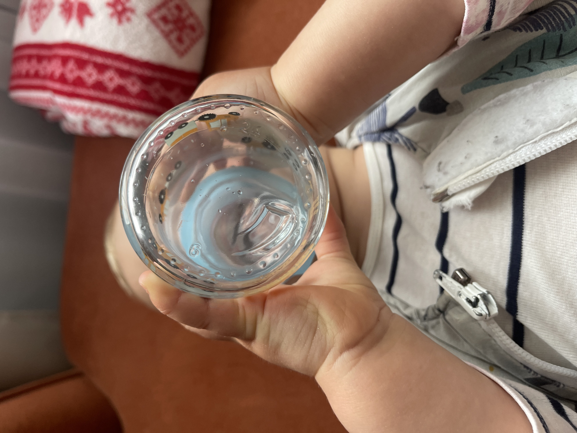 A small child holds a clear glass of water with a blue silicone sippy cup insert. The child is wearing a white romper with thin navy blue stripes. A white towel with a red patterned border is visible in the background on what appears to be an orange surface. The location is unknown and the image provides no information on its origins in Sofia, Bulgaria.