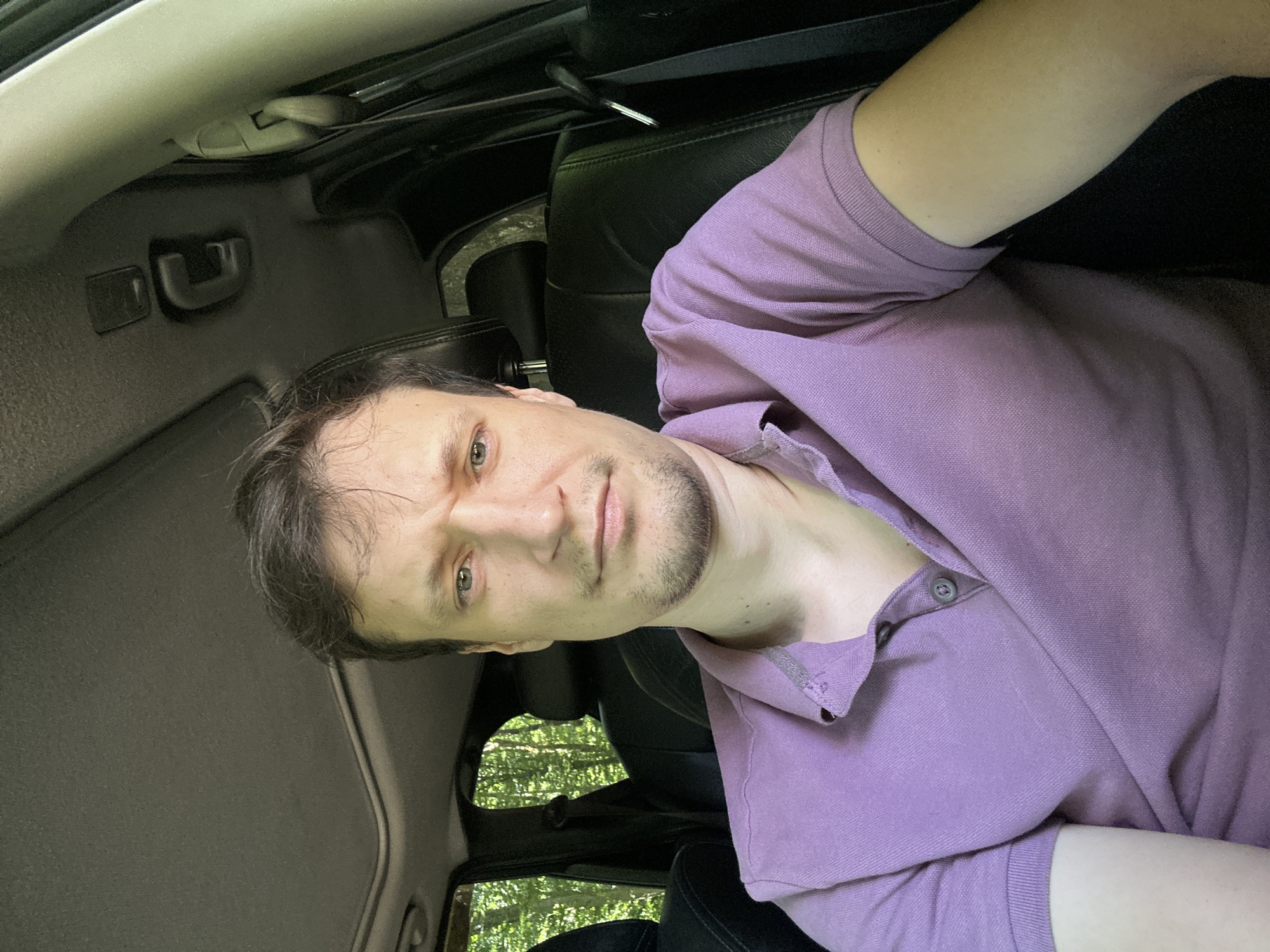 A man is sitting in the driver's seat of a car. He has short, dark brown hair and a light complexion. He is wearing a purple polo shirt. His expression is neutral, with his brow slightly furrowed. The car's interior is dark, with leather seats visible. Outside the car, trees are visible through the windows, suggesting a wooded area. The image appears to be taken in Shatava, Ukraine.