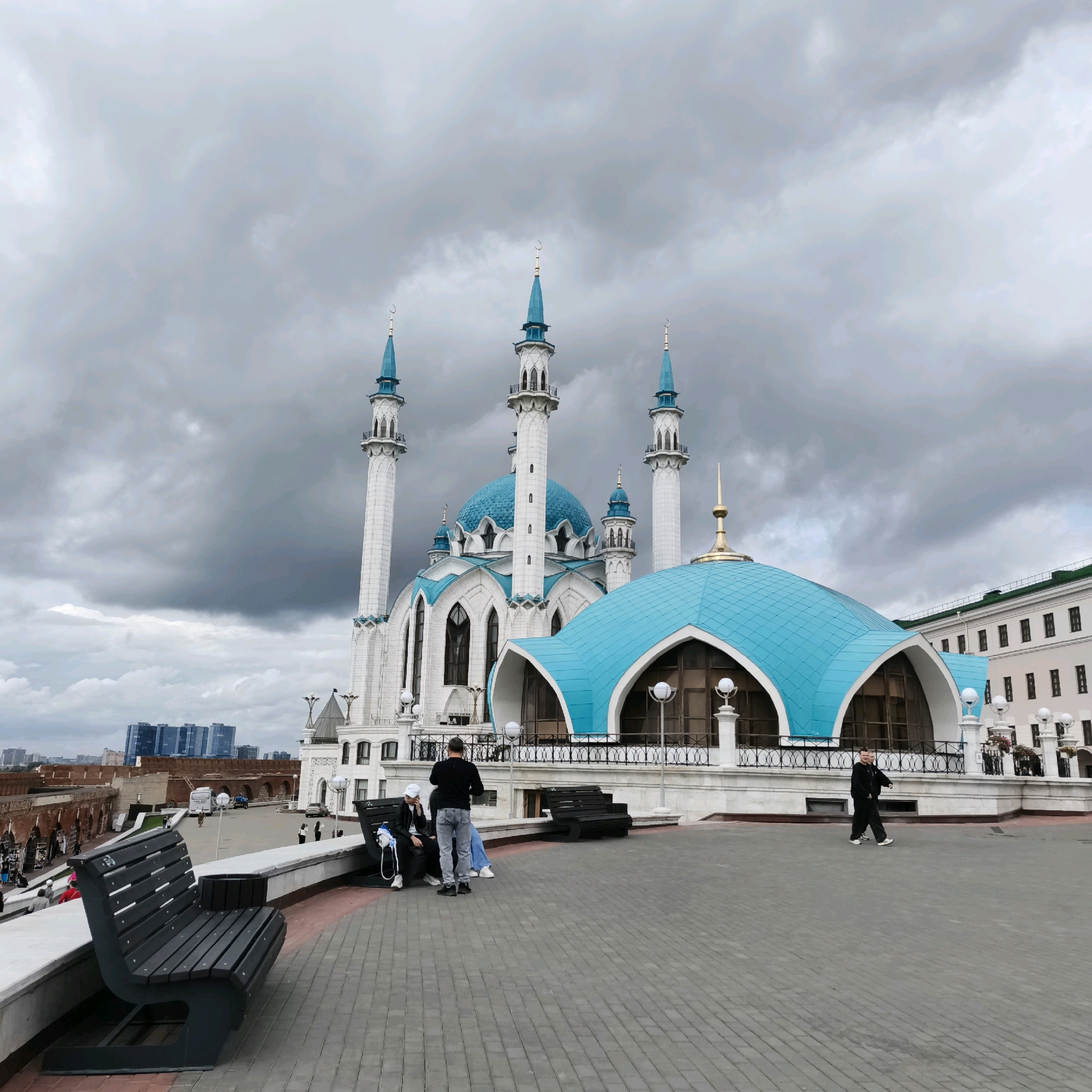 This is an outdoor, eye-level, medium shot photograph of the Kul Sharif Mosque in Kazan, Russia, under a cloudy sky. The mosque is the central subject, distinguished by its white minarets topped with blue spires and a large, turquoise dome. The architecture is ornate, with decorative elements and arched windows visible. In the foreground, a dark-colored park bench with a modern design is positioned on a paved walkway. Two individuals are seated on the bench, facing away from the camera. Another individual stands to the left of the bench, facing the mosque and appears to be taking a picture. Further to the right, two people walk away from the camera. In the background, a section of a fortress wall, also made of brick, can be seen. Beyond the fortress, several modern buildings with glass facades rise into the distance. The overall atmosphere is subdued due to the overcast weather.