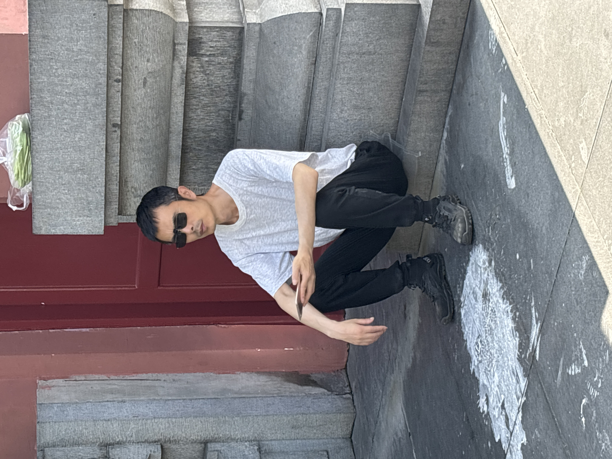 A man wearing sunglasses, a white t-shirt, and black pants sits on a stone step outdoors. He holds a mobile phone in his left hand. A plastic bag containing green leafy vegetables rests on the stone ledge above him. The ground in front of him is speckled with a white substance. A red door is visible to the left of the man. The architecture suggests the location is likely in Yuzhong District, China.