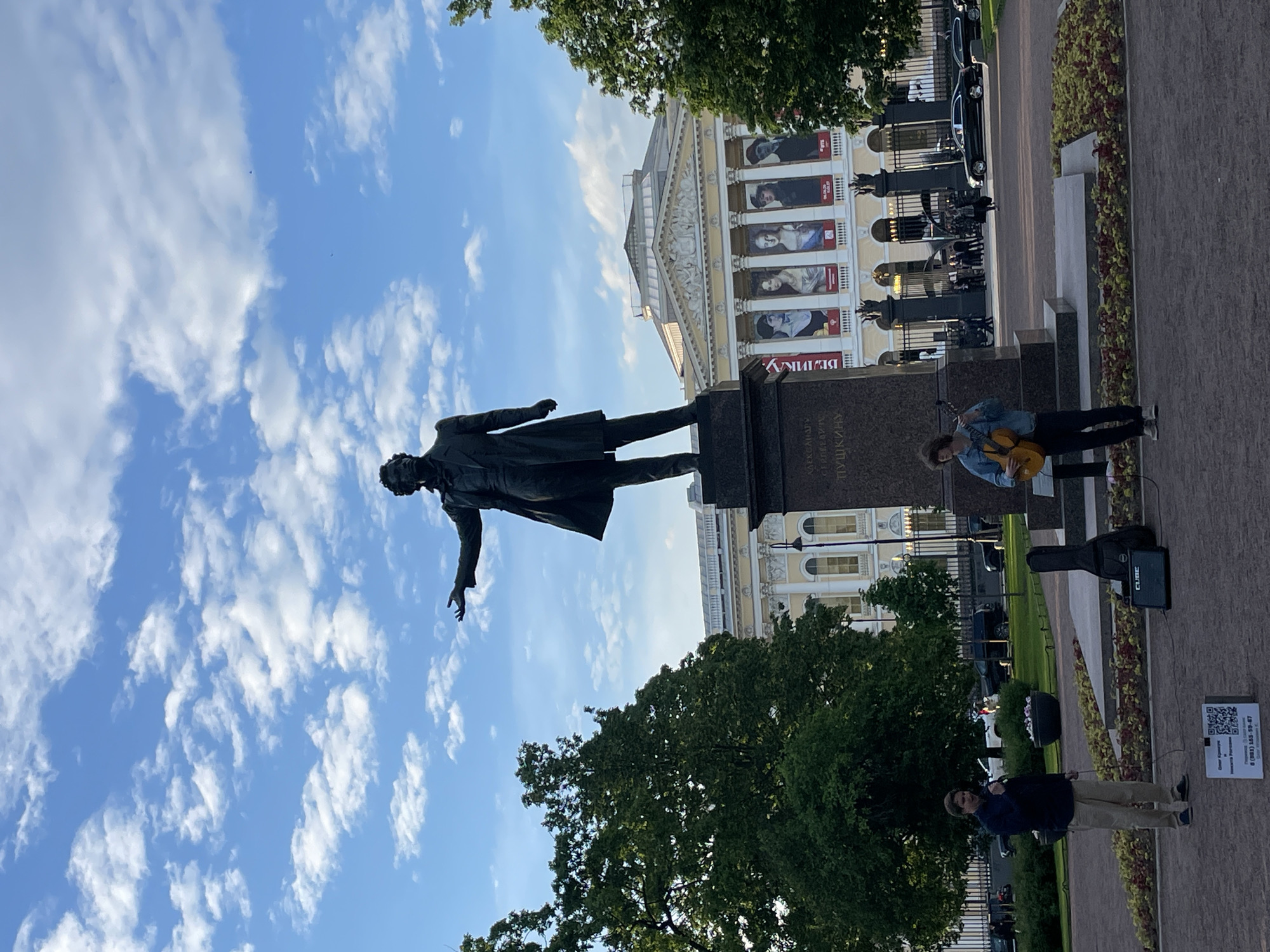 This image captures a vibrant street scene in Saint Petersburg, Russia, centered around the iconic Pushkin Monument. The bronze statue, depicting a distinguished man in a long coat with one arm extended, stands atop a dark stone pedestal. An inscription in Cyrillic on the pedestal reads "АЛЕКСАНДРУ СЕРГЕЕВИЧУ ПУШКИНУ" (To Alexander Sergeevich Pushkin).
In front of the monument, a street musician performs, playing an acoustic guitar. He wears a denim jacket and a hat, and stands near an open guitar case and a black portable amplifier, suggesting he is busking for donations. Further to the right, another person in a dark jacket and light pants stands near a white sign.
The background features a grand, classical-style building, likely the Alexandrinsky Theatre, with a yellowish facade, numerous arched windows, columns, and decorative moldings. Large banners featuring portraits, possibly of actors or historical figures, hang from the building's exterior. Part of the Cyrillic word "ВЕЛИКУ" (Great) is visible on one banner. Lush green trees frame parts of the scene, and a decorative fence runs behind the monument, separating the public space from a street with parked cars.
The time of day appears to be sunny daytime, with a bright blue sky dotted with scattered white cumulus clouds. The overall atmosphere is lively and pleasant. The white sign on the ground near the second person displays a QR code, alongside Cyrillic text that translates to "Fundraising for publication and help Ruslan," and includes a phone number: "8 (981) 185-59-87."
