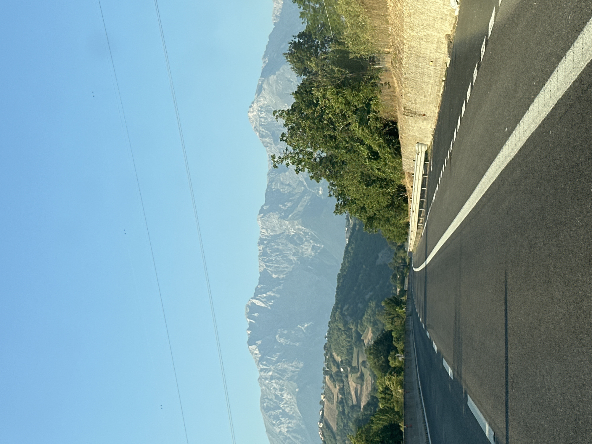 The image depicts a highway leading towards a mountain range. The road has clear white lane markings and guardrails. The surrounding environment is hilly, with trees and some scattered buildings visible on the slopes. Power lines are present in the sky, and a few birds are flying. The mountains in the background are rugged and appear to be covered in patches of rock and vegetation. The sky is clear and blue. This scene is consistent with the landscape found in and around Colledara, Italy.