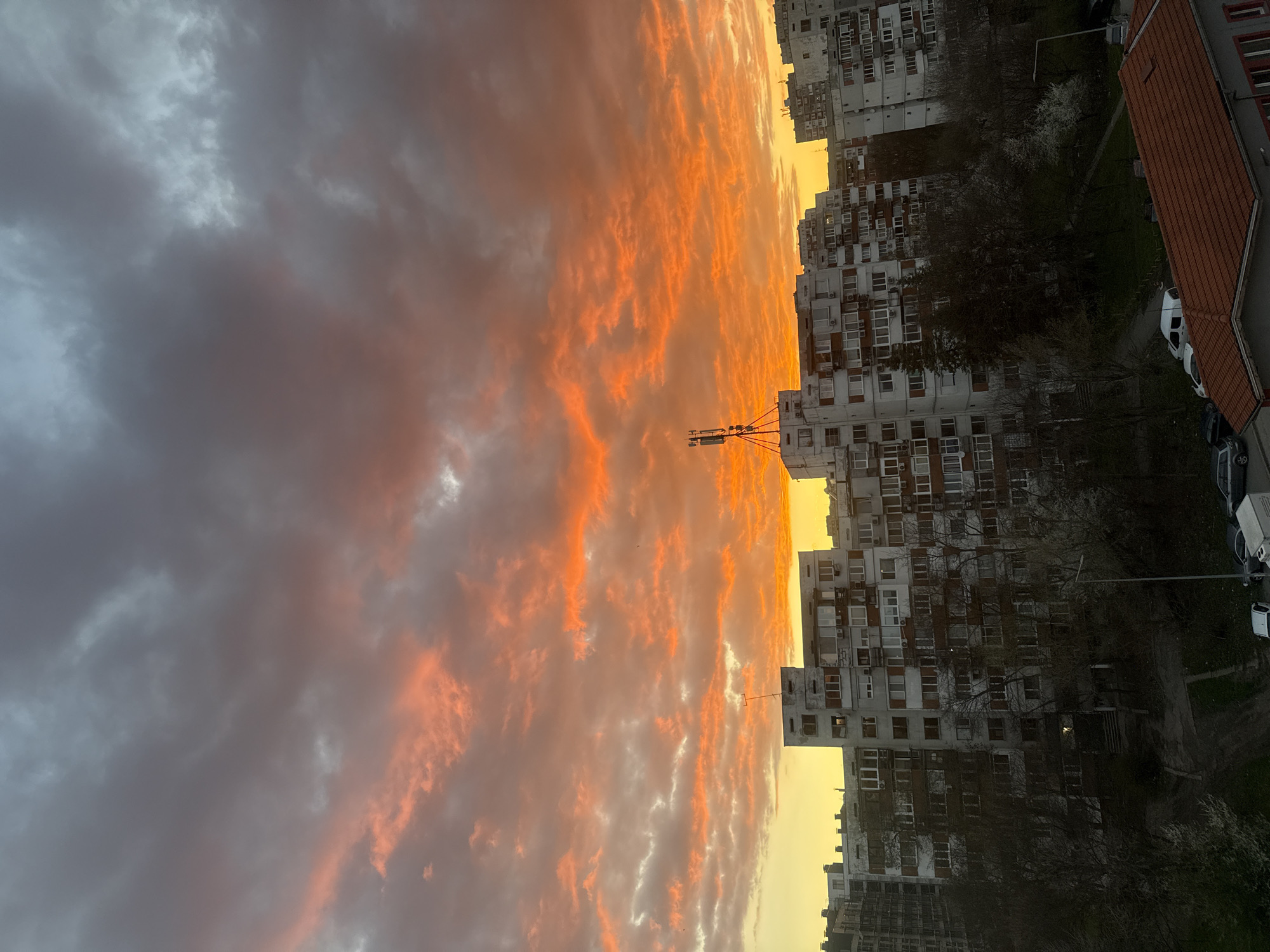 The image shows a sunset in Sofia, Bulgaria, with high-rise apartment buildings lining the horizon. The sky is filled with orange and grey clouds. The buildings appear to be residential apartments with many windows and balconies. A utility pole with attachments stands out against the sky. In the lower right corner, there's a view of rooftops, trees, and parked cars. The photograph is taken from a high vantage point, possibly an upper-floor apartment or building.