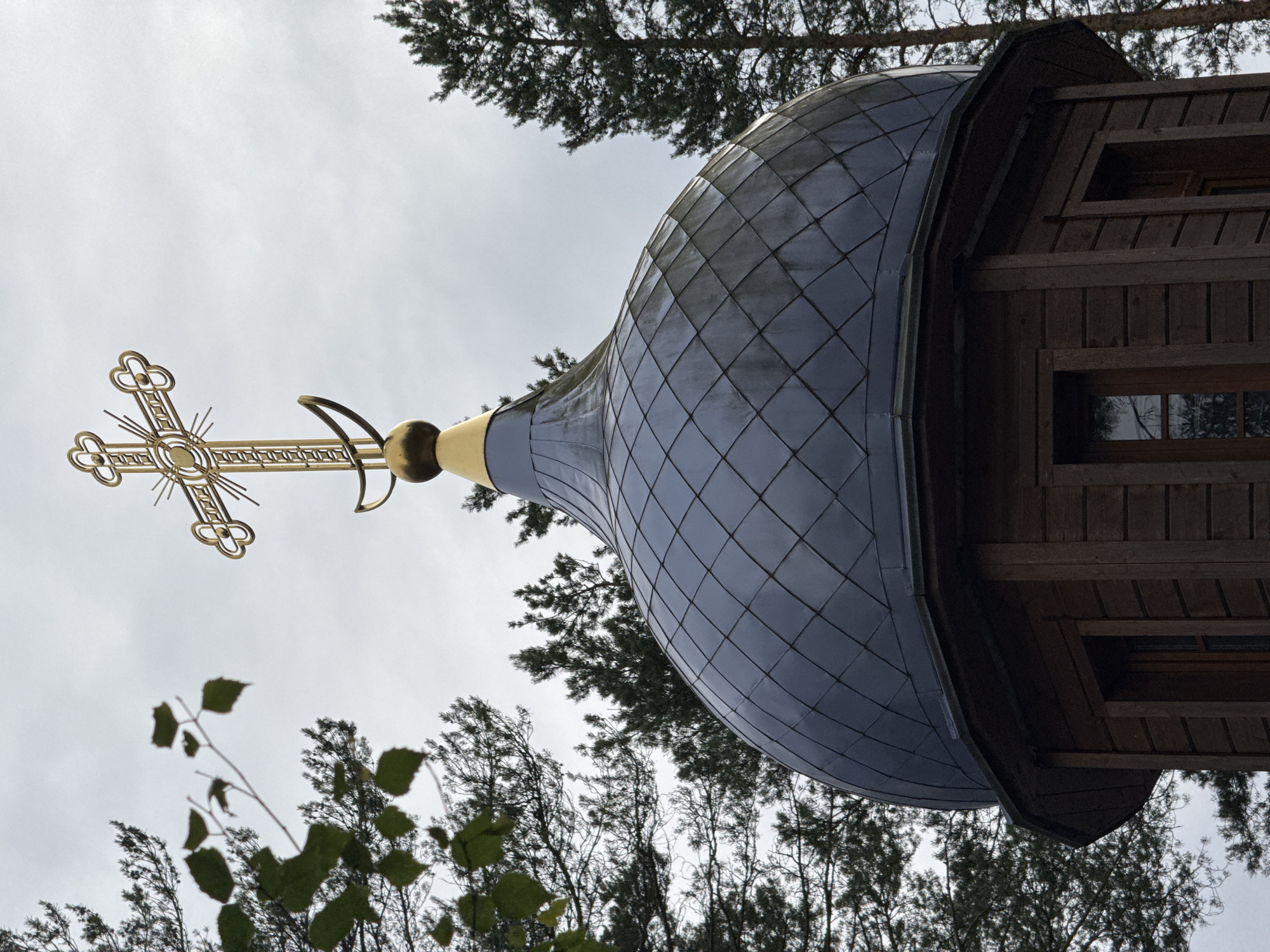 A close-up, eye-level, outdoor shot shows the top of a wooden Orthodox church, likely located in Nenyamyaki, Russia. The dome is covered in dark, diamond-shaped metal tiles, and it is topped with a golden cross and crescent moon. Tall, dark green pine trees surround the church. The sky is overcast and a pale gray. In the foreground to the left, a branch with green leaves hangs into the frame.
