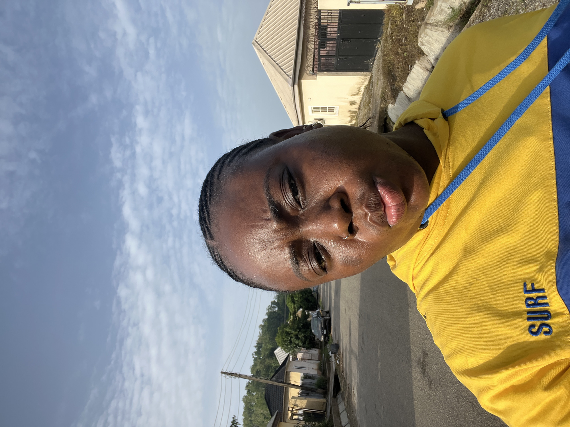A close-up photograph shows a person in a yellow and blue hooded sweatshirt standing on a paved road. The word "SURF" is visible on the sleeve. The person has dark skin, braided hair, and a nose ring. The background reveals a partly cloudy sky and residential buildings. The location appears to be a street in a residential area, likely in Abuja, Nigeria, given the landscape and architectural style of the buildings. A car is parked further down the street. Power lines run along the side of the road.
