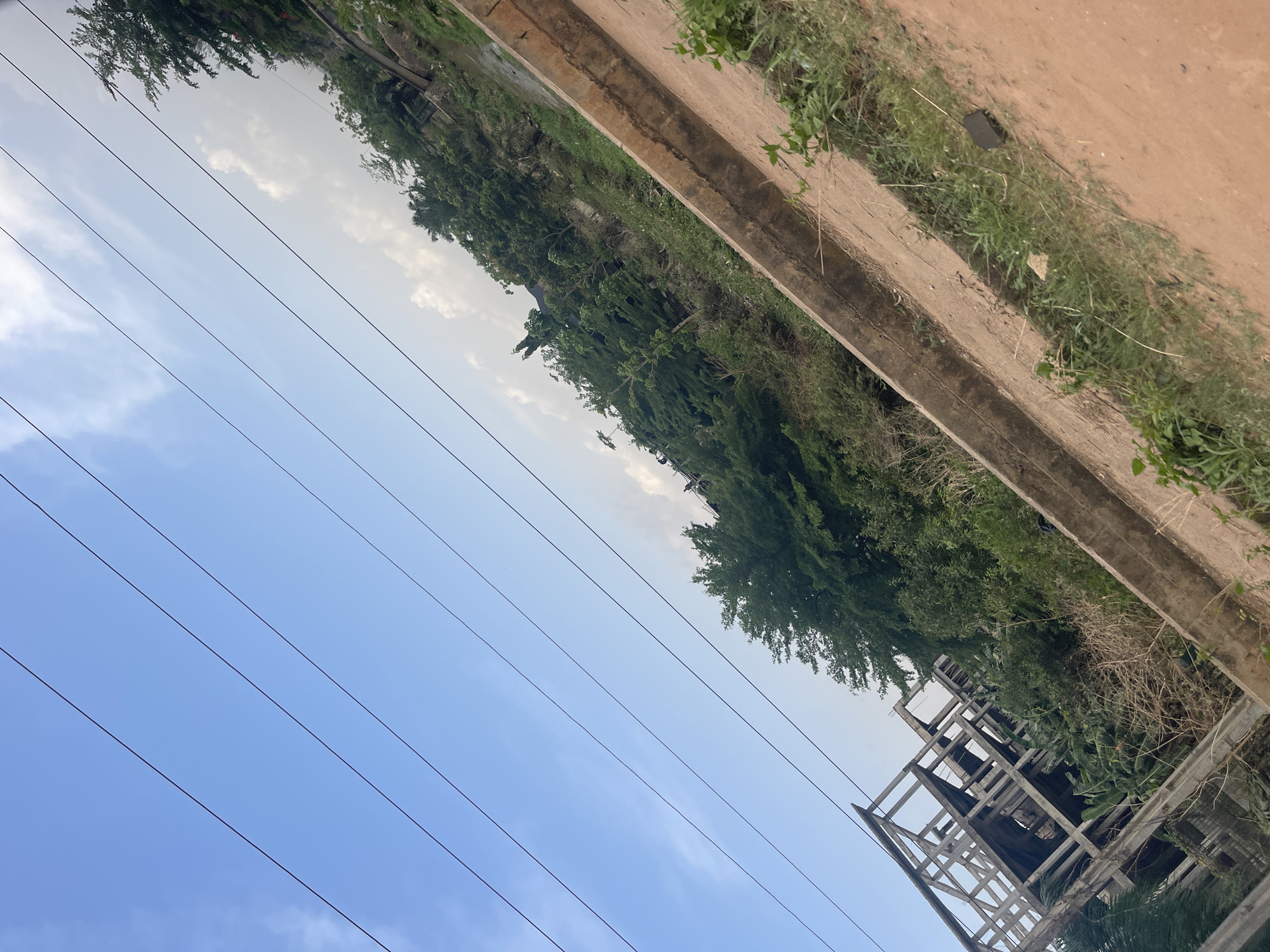The image captures a daytime landscape scene, likely in Gwarinpa, Nigeria, characterized by natural elements and emerging infrastructure. The upper half of the frame is dominated by a clear blue sky with scattered white clouds. Multiple black power lines stretch diagonally across the sky from left to right, suggesting an urban or suburban setting with electricity access.
Below the sky and power lines, a dense line of green trees and various types of vegetation forms a verdant backdrop, extending horizontally across the middle of the image. Some areas within the foliage show drier, brownish plants. Parallel to the vegetation, a light brown dirt path or unpaved road is visible, flanked by a concrete strip or gutter that separates it from the greenery. This concrete structure appears as a low barrier or drainage channel.
On the right side of the image, amidst the vegetation and extending upwards, is the skeletal framework of a multi-story concrete building, indicating ongoing construction or an unfinished structure. In a cropped detail, a small, dark, rectangular object can be seen on the grassy area adjacent to the dirt path.
The scene is static, with no discernible human activity or moving objects. The bright natural light indicates it is either morning or afternoon with fair weather. There is no visible text in the image.