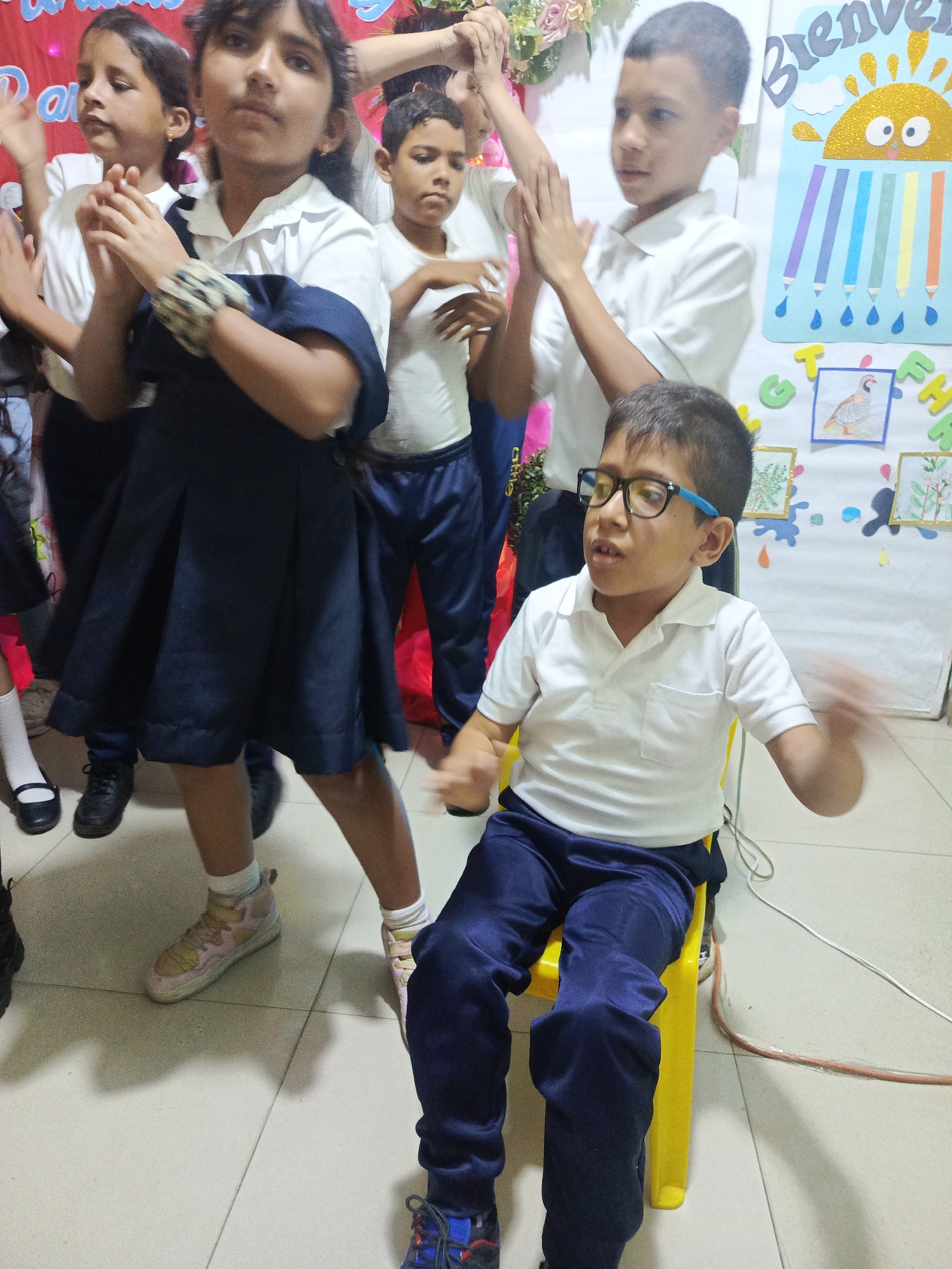 A group of children in school uniforms are gathered in what appears to be a classroom in Juan Griego, Venezuela. The children are mostly standing and clapping, while one child is seated in a yellow plastic chair, also clapping. A banner or bulletin board with the word "Bienve..." is visible behind them, along with other decorations featuring a sun, pencils, and animal pictures. The floor is tiled and the environment appears to be well-lit.