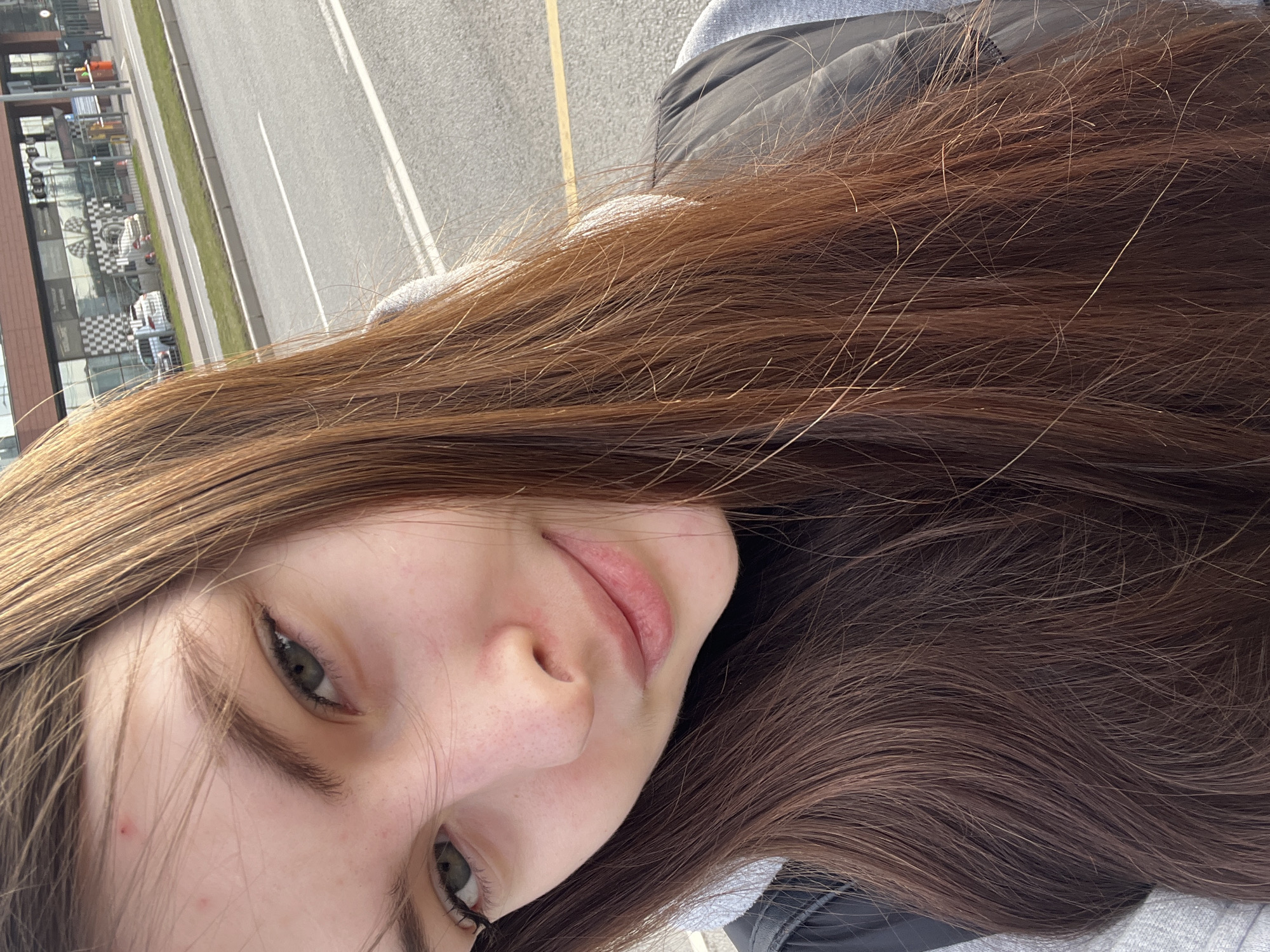 A young woman is photographed in a self-portrait style. She has long, brown hair and fair skin. She has green eyes and light pink lips. She is wearing a gray hooded sweatshirt and a black jacket. The background shows a modern building with glass windows and a parking lot. The building appears to have a black and white checkered pattern on some windows. A gray road with white lane markings and a yellow center line is visible. Based on visual cues like architectural style and road markings, the location may be in Saint Petersburg, Russia.