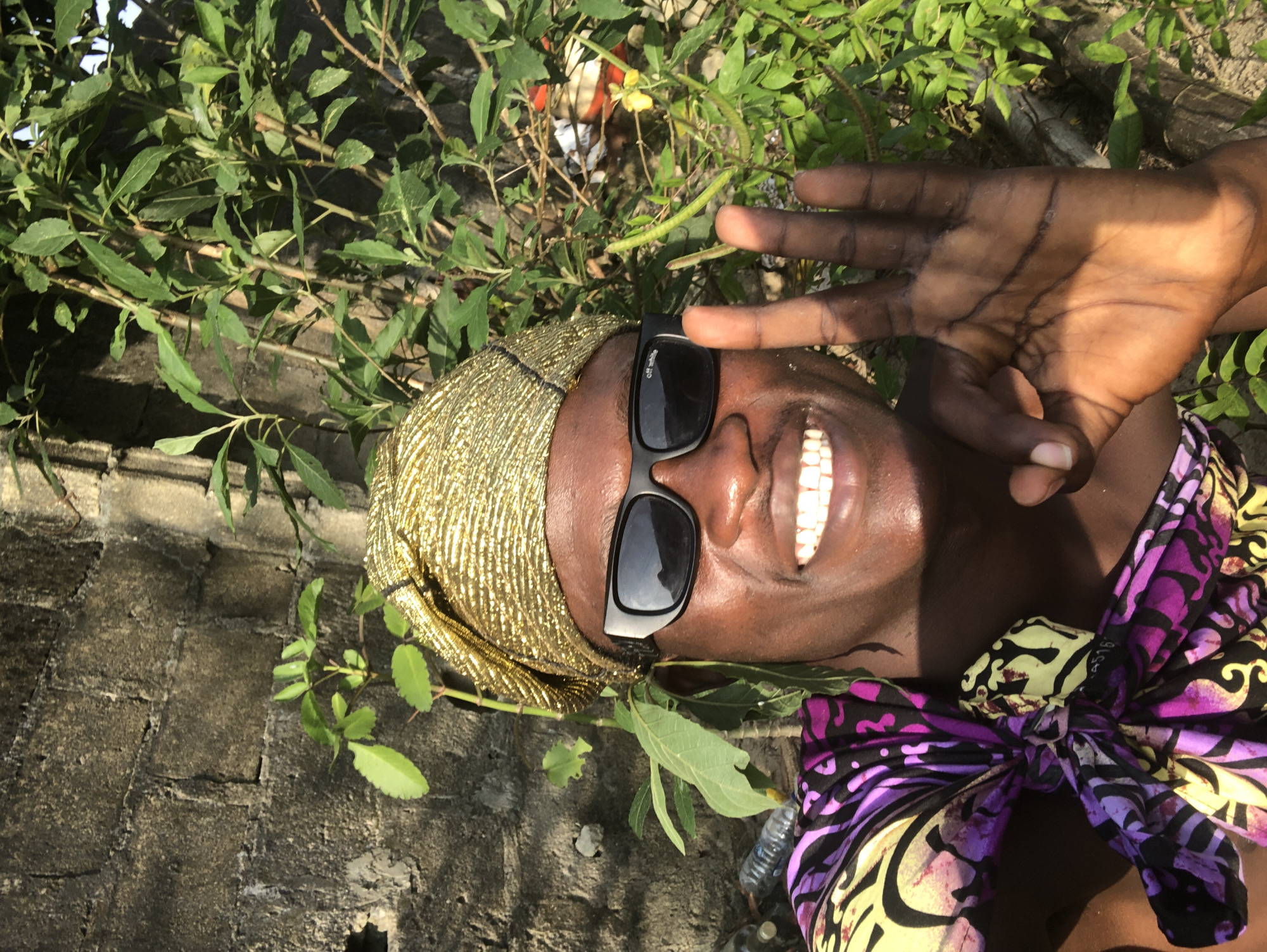 A close-up selfie shows a person smiling and holding up their right hand in an "okay" gesture. They are wearing a gold headwrap, black sunglasses, and a colorful patterned top. The person's skin tone is dark brown. The background consists of a textured brick wall and lush green foliage. Some plastic water bottles are visible at the bottom left of the image. The lighting suggests an outdoor setting, likely during the day.