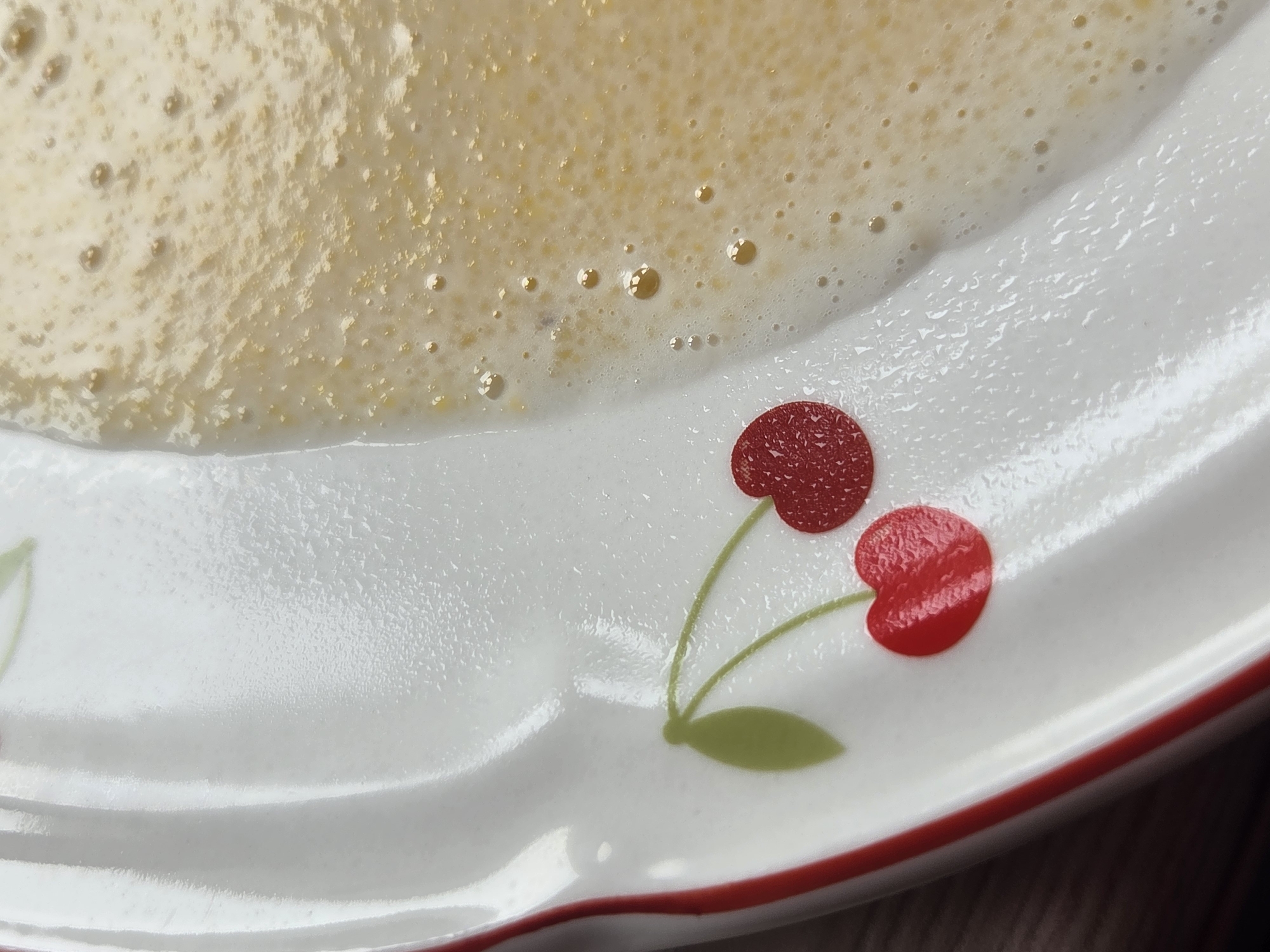 The image shows a close-up view of a white ceramic plate with a red cherry design. A portion of the plate is covered with a foamy, light brown liquid that appears to be some sort of batter or sauce. The liquid has small bubbles and a slightly textured surface. The plate has a red rim and a decorative pattern of cherries and leaves. The lighting suggests an indoor setting. The location is the city of Восход Снт (фролово), Russia.