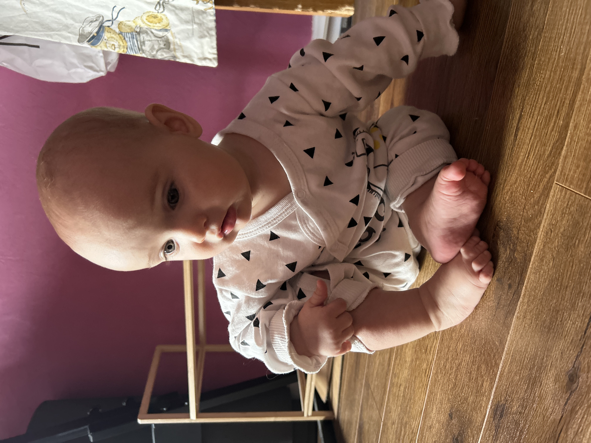 A baby, wearing a white outfit with black triangle patterns, is sitting on a wooden floor. The baby is holding its foot with its hand. A pink wall is visible in the background, along with a wooden structure. A white cloth with cartoon-like illustrations hangs from a nearby object. There is no indication of this image being in Moscow, Russia.