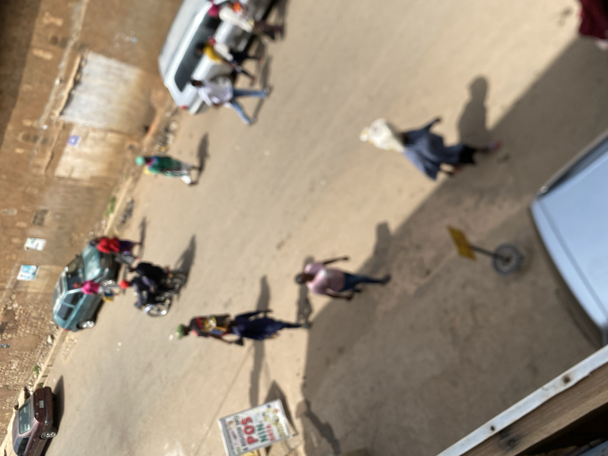 The image, taken from an elevated position, shows a street scene in Bukuru, Nigeria. Several people are visible, walking or standing in various locations. Several vehicles are parked on the street. There is signage visible on the street.