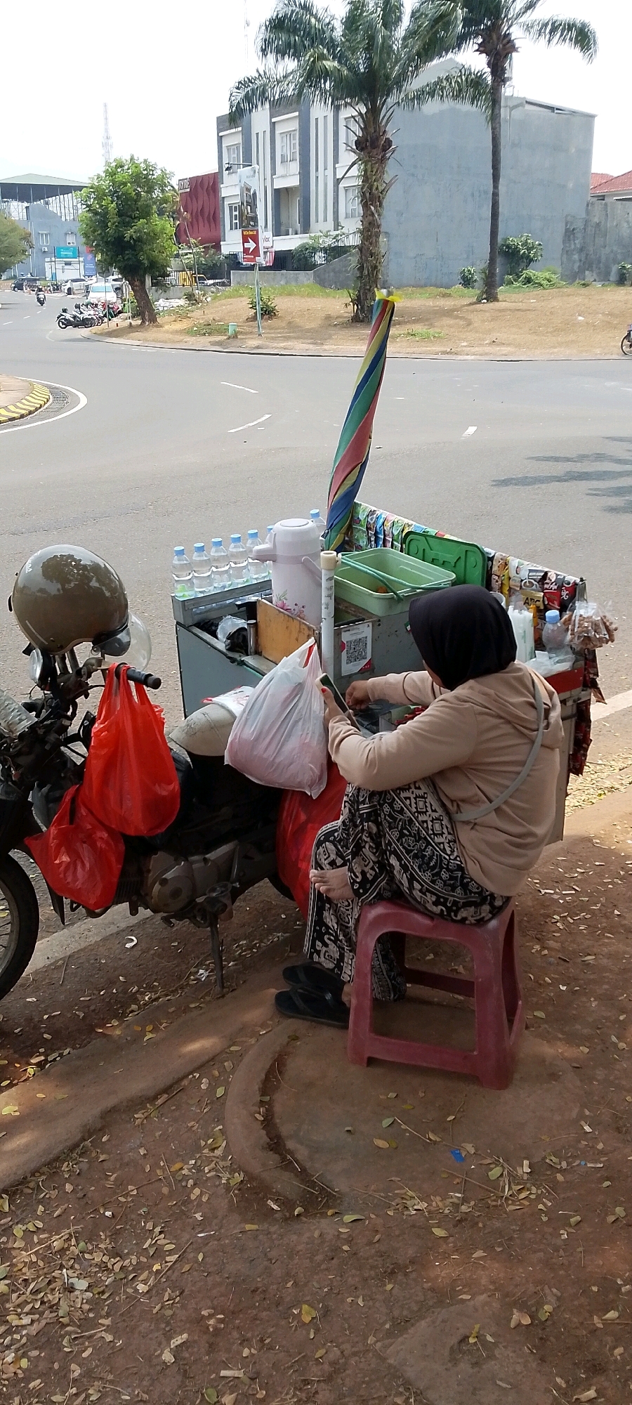In Gunung Putri, Indonesia, a person is sitting on a red stool next to a motorcycle and a street food cart. The cart is laden with bottled water, a thermos, and various food items. The person, wearing a hijab and a light-colored hoodie, is holding a mobile phone and appears to be engaged with it. Red and white plastic bags hang from the motorcycle, and a colorful umbrella is attached to the cart. The scene unfolds on a roadside, with a paved street and a building in the background. Fallen leaves litter the ground, suggesting an outdoor setting.