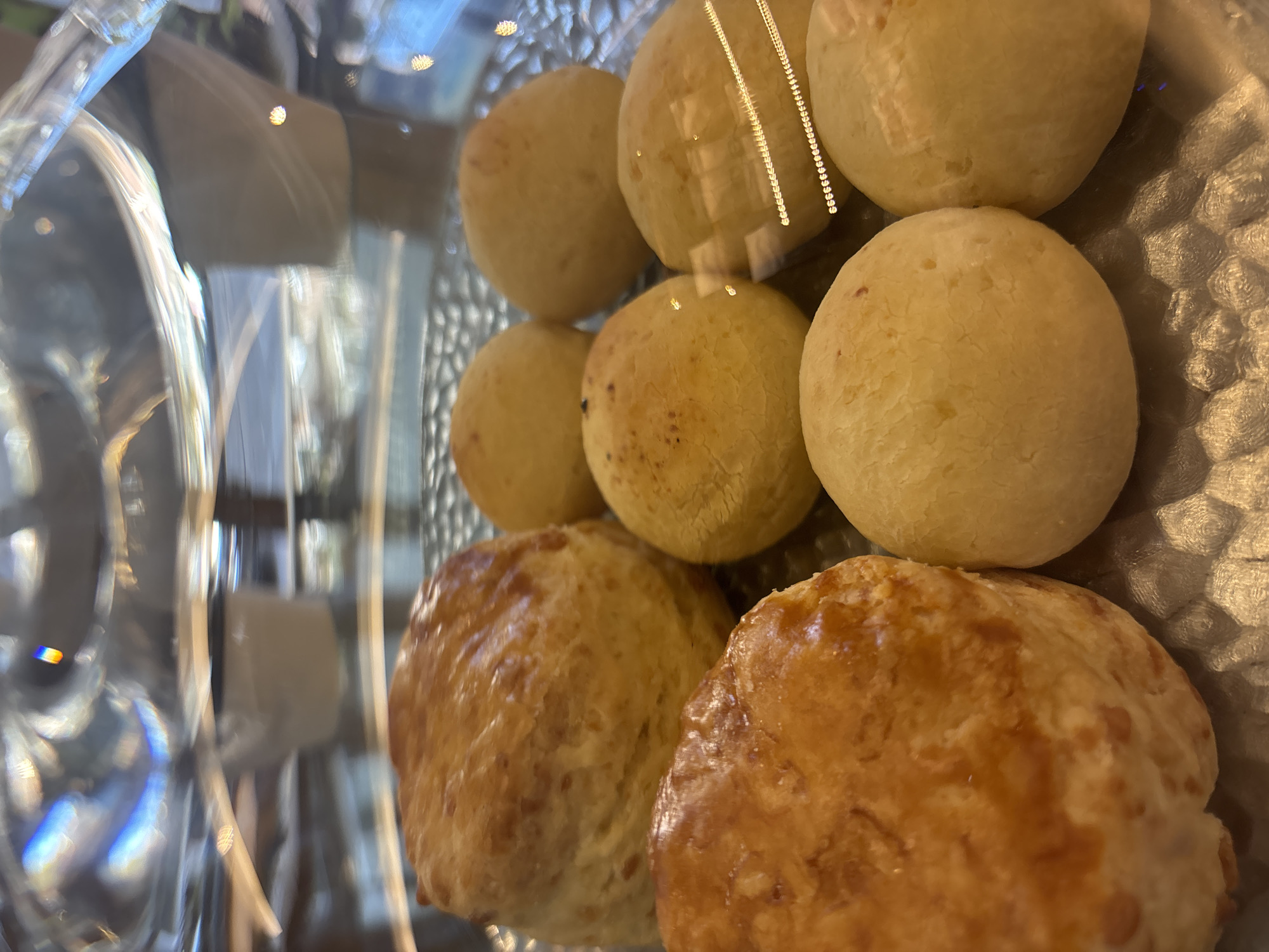 The image shows an assortment of baked goods displayed inside a glass case. At the top, a row of small, round, pale-yellow buns are arranged, reflecting light. Beneath them, a second layer consists of similarly shaped buns, some with slight browning. In the foreground, two larger, golden-brown, slightly cracked buns are visible. The glass case distorts the objects visible through it. The location is unknown.
