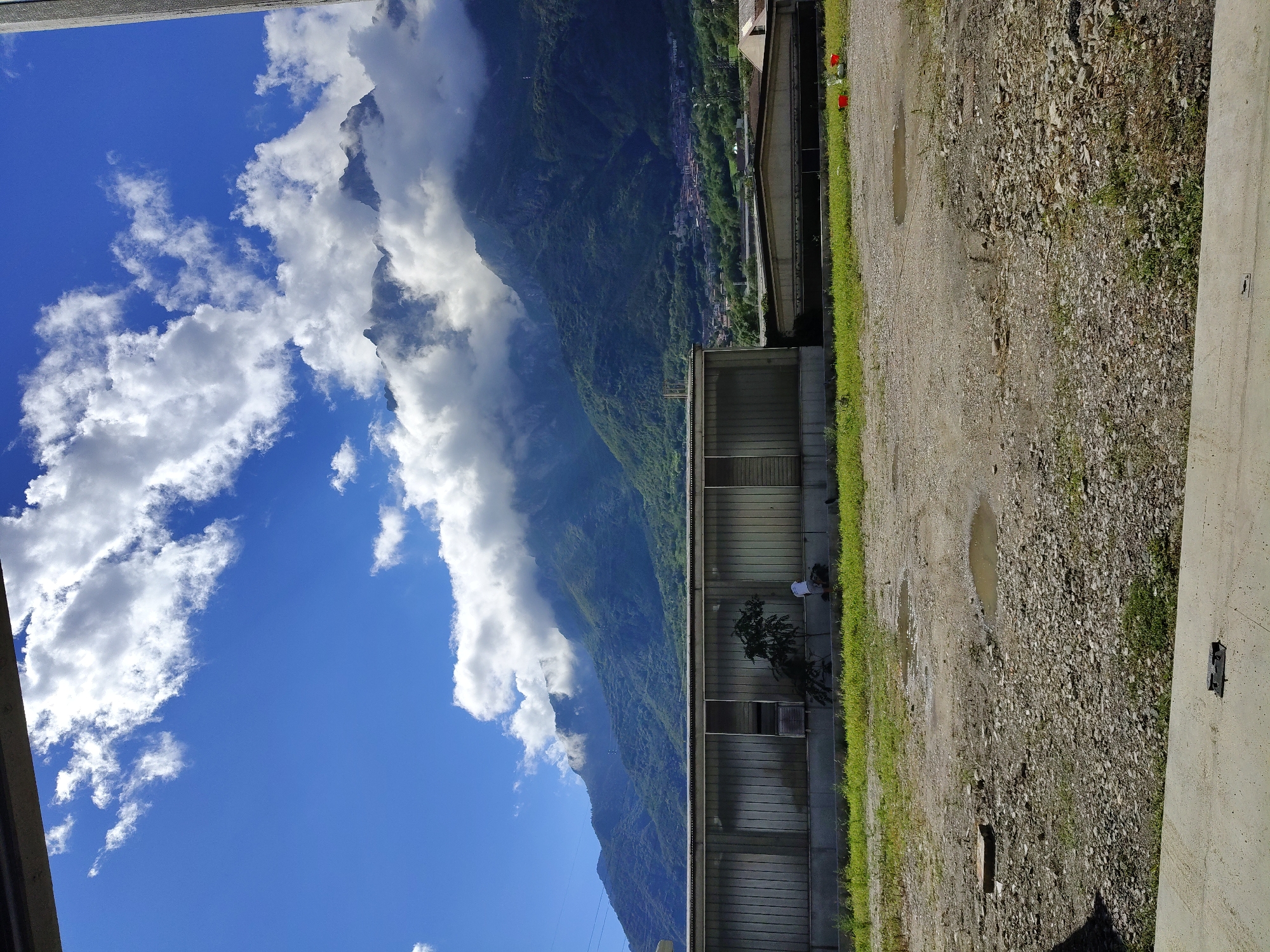 The image depicts a clear, bright day with a blue sky filled with fluffy white clouds. In the foreground, a gravel and dirt lot is visible, with patches of grass and several puddles. A long, metallic-sided building with glass panels runs horizontally across the middle ground. To the right of this building, a smaller structure with a pitched roof is visible, and two red buckets and some green plastic bags are on the ground nearby. Further in the background, a range of lush green mountains rises, with a small town nestled at their base. The peaks of the mountains are partially obscured by the clouds. A person in a white shirt is seen near the metallic building, tending to a small tree.