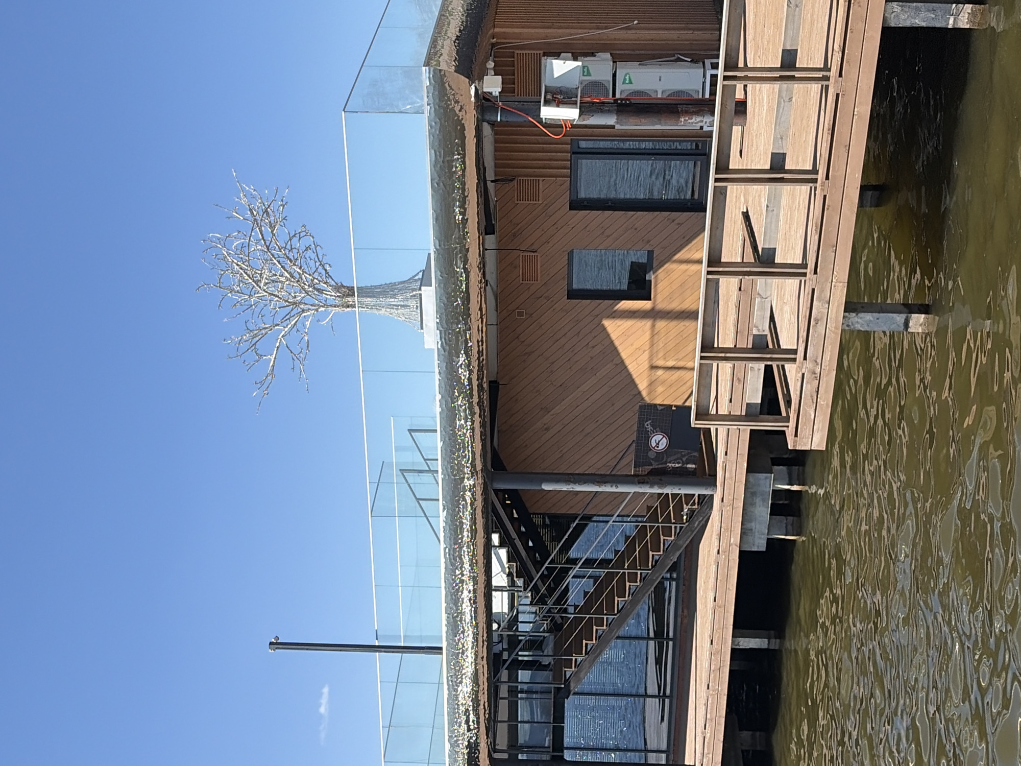 The image shows a building on stilts in Votkinsk, Russia. The building has a wooden facade and a glass-enclosed rooftop with a decorative tree-like structure. There are stairs leading up to the building and a wooden ladder extending into the water. Air conditioning units are visible on the building's exterior. The water surrounding the building appears calm. The sky is clear and blue.