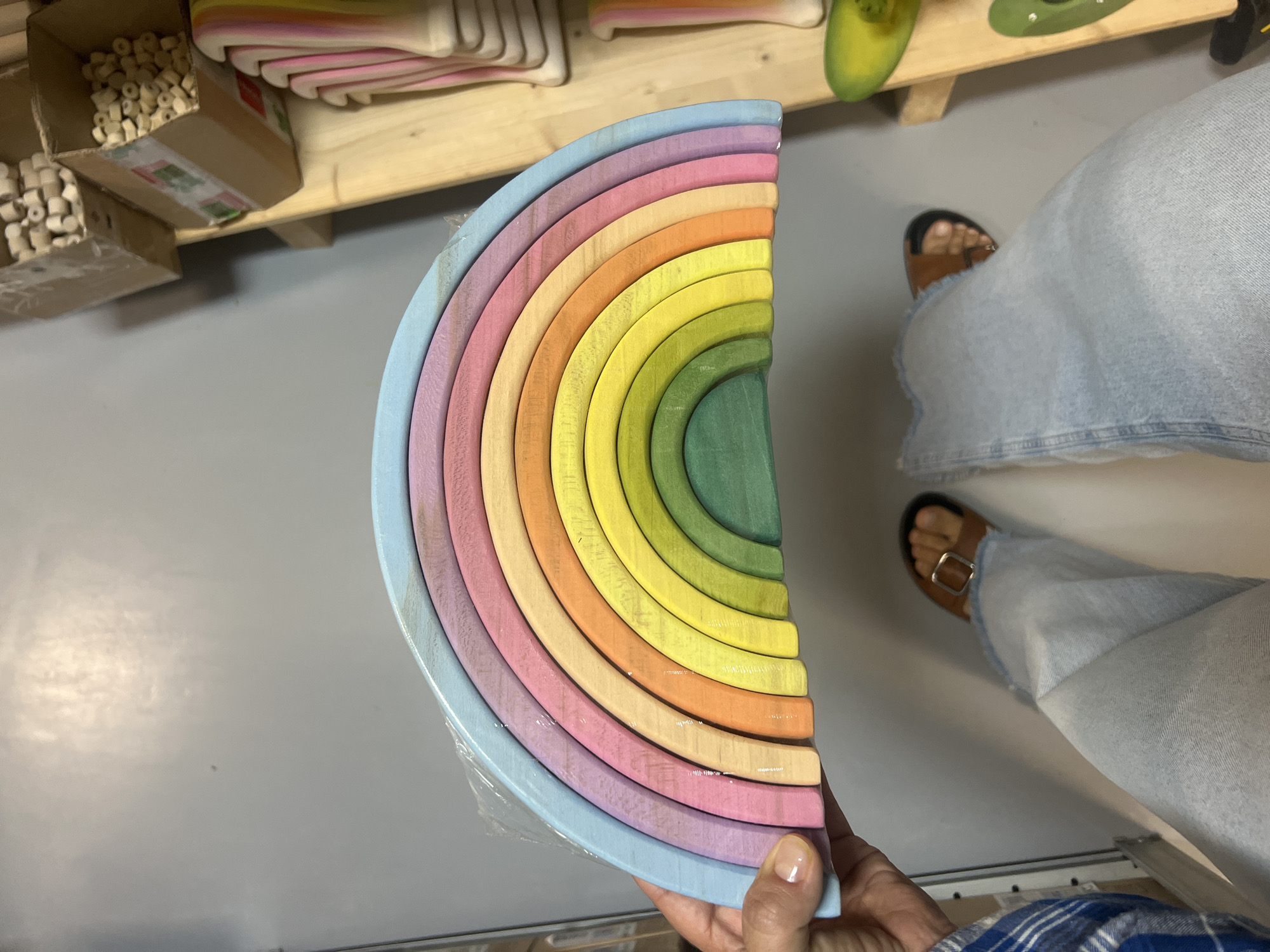 A person is holding a colorful wooden rainbow toy. The toy is made of curved wooden pieces in various pastel colors, including blue, purple, pink, orange, yellow, and green. The person's hand, with painted fingernails, is visible holding the lower portion of the toy. The person is wearing light blue denim jeans and brown sandals. The setting appears to be indoors, possibly a workshop or store, with wooden shelves and boxes visible in the background. Some of the boxes contain small wooden beads. The lighting suggests it is daytime. The overall mood is casual and perhaps indicative of a retail environment or hobby space.