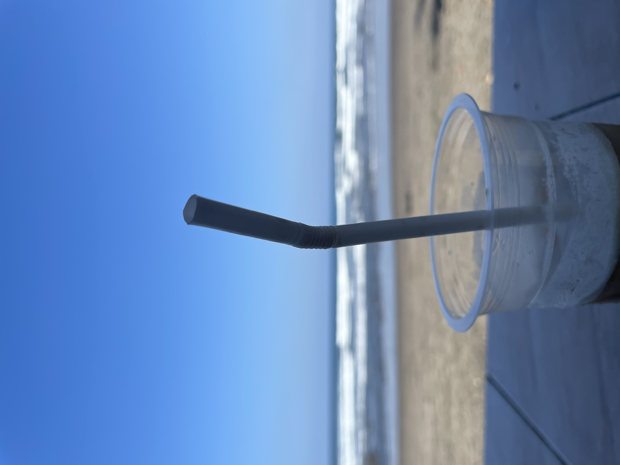 A plastic cup with a straw sits on a dark, reflective surface, possibly a table. The cup contains a frothy beverage. In the background, a beach stretches out towards the ocean, with waves gently rolling onto the shore. The sky above is a clear, bright blue. The scene is likely situated in Pekutatan, Indonesia, given the tropical beach setting.