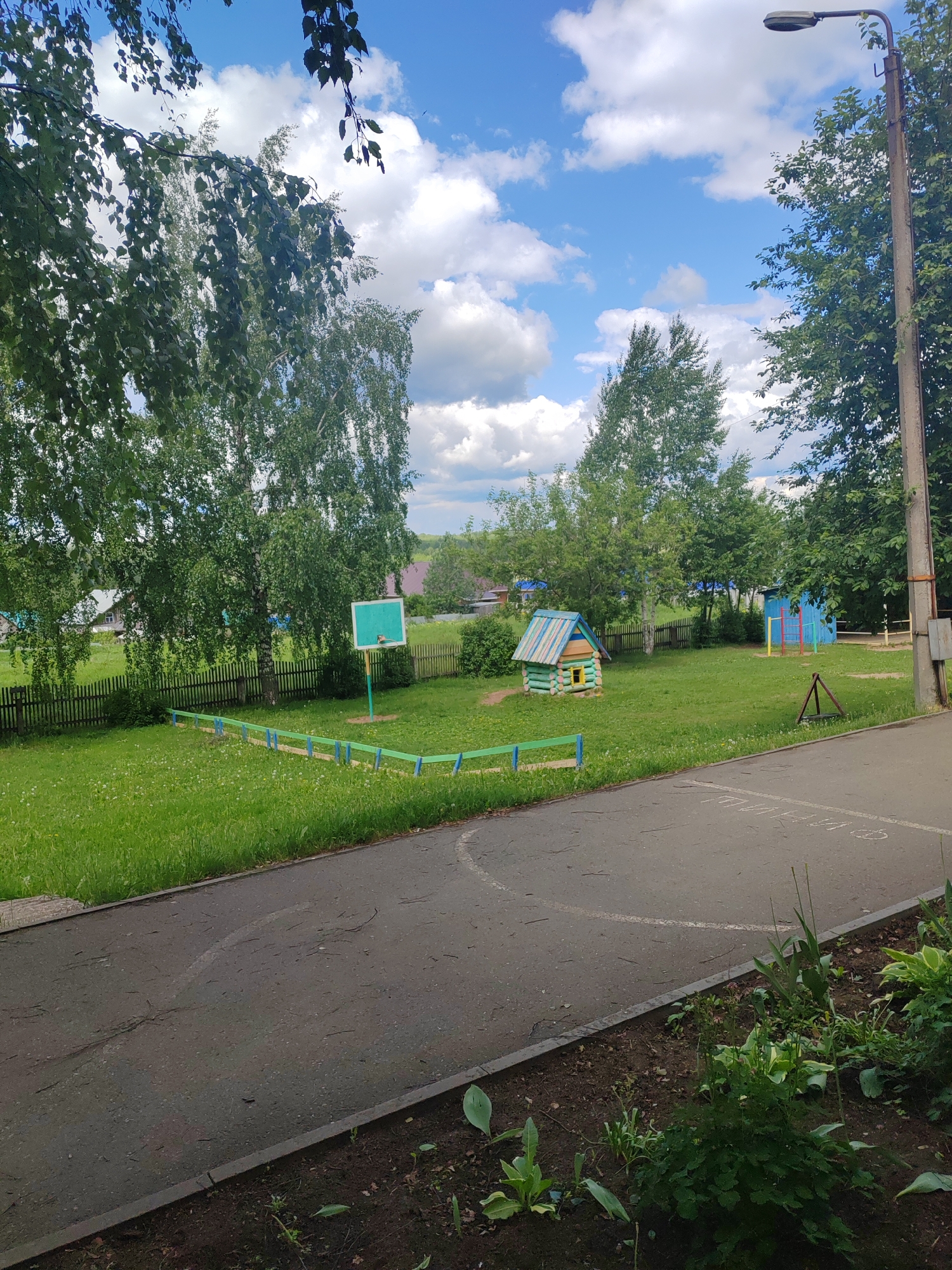 The image captures a tranquil scene in an urban park within the city of Unknown. The park appears to be a recreational area with play structures and elements suitable for children.
The environment is bathed in daylight, under a blue sky dotted with fluffy white clouds. Lush green trees frame the background. A small, wooden playhouse with a multi-colored roof and a tiny window can be seen in the center of the scene. Other play structures, including a basketball hoop and a blue painted structure with a red ladder, are visible in the background. A green and blue painted fence section is visible.
A grey asphalt pathway extends into the foreground from the lower right corner. Alongside the path is a patch of cultivated soil with plants. A light post stands tall, and the ground cover is a vibrant mix of grass and wildflowers.
The scene suggests a safe, quiet neighborhood suitable for families with young children.