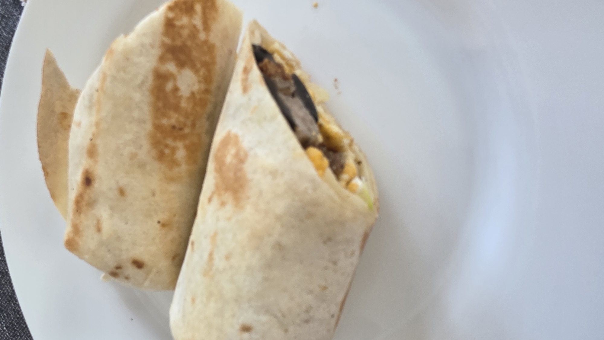 A close-up shot of two burritos cut in half on a white plate. The burritos appear to be filled with eggs, meat, and vegetables. The background is a plain white plate with a small portion of a gray table visible on the left edge. The scene is indoors with natural lighting. Location details (Blagoevgrad, Bulgaria) cannot be determined from the provided image.